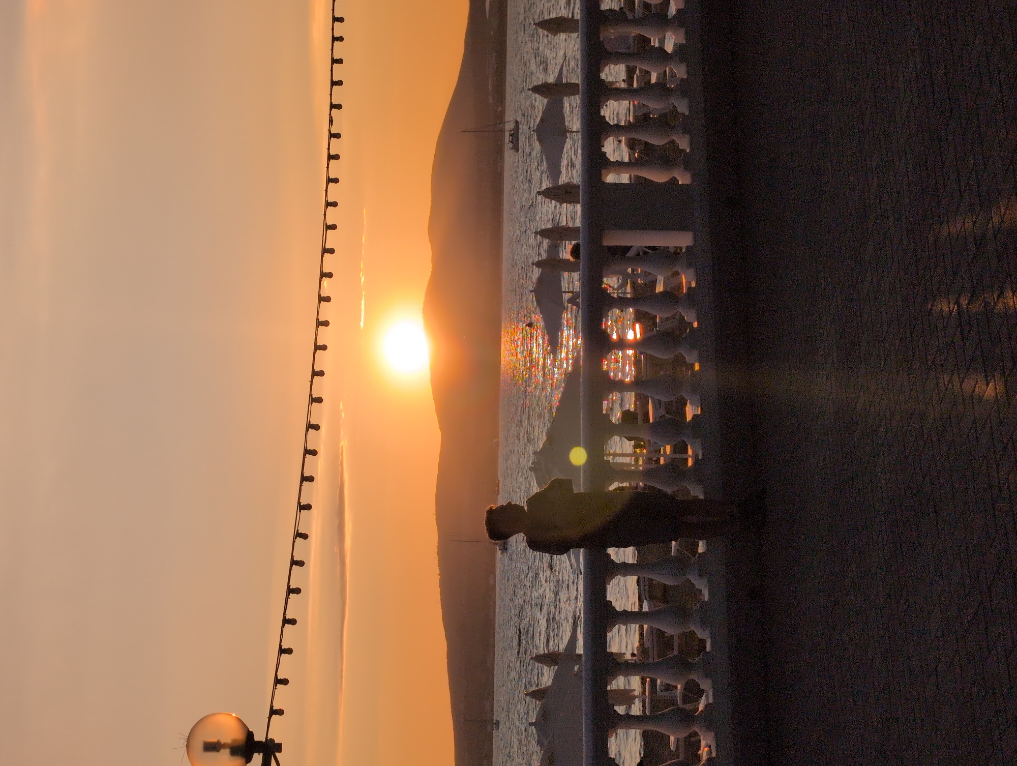 A person stands on a paved walkway overlooking a body of water at sunset. The sun is low on the horizon, casting a warm orange glow across the sky and reflecting on the water. The person, silhouetted against the sunset, is facing away from the camera, looking towards the sea. In the background, there are hills and sailboats on the water. A white railing with decorative posts separates the walkway from the water. A string of lights hangs overhead, illuminated by the sunset. The overall scene is peaceful and serene.