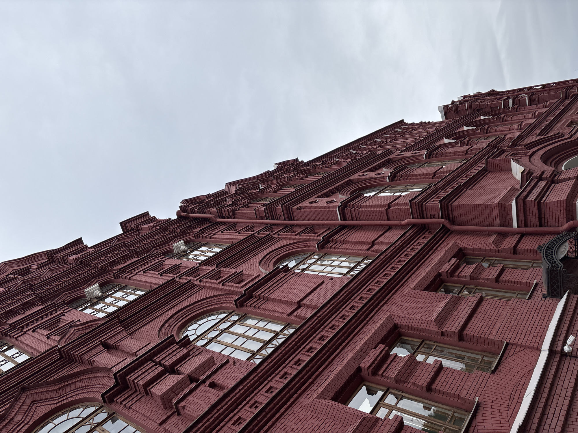 The image shows a low-angle view of a red brick building against a cloudy sky. The building's facade is highly detailed with arched windows and intricate brickwork. The location is likely in Moscow, Russia.