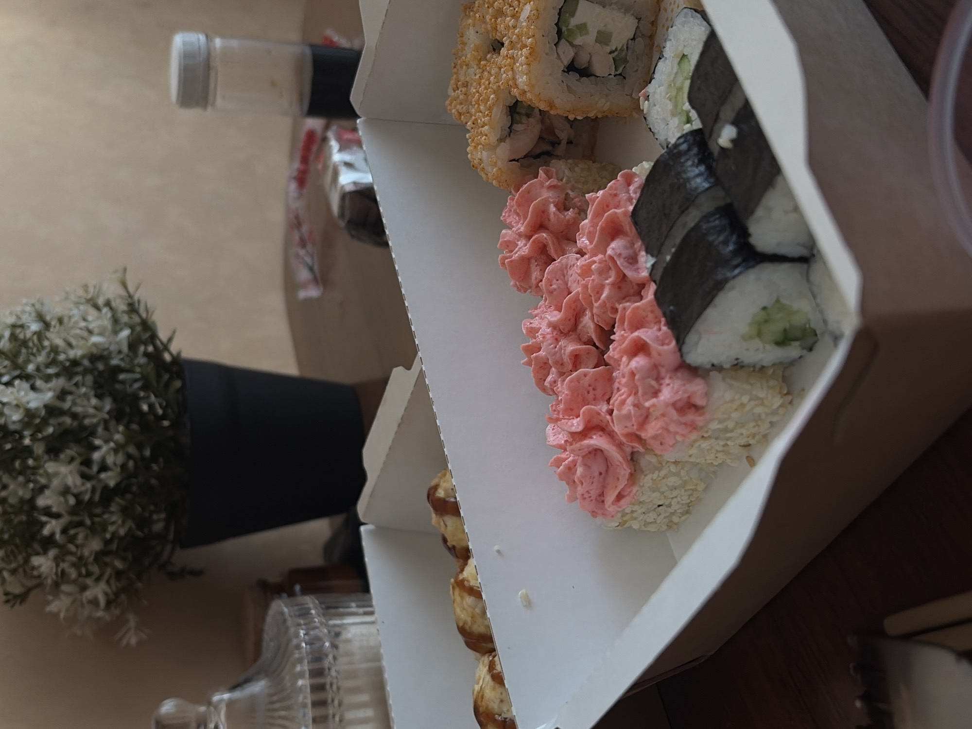 The image shows a variety of sushi rolls in a white cardboard takeout container. Types visible include rolls covered in sesame seeds with a pink topping, rolls wrapped in nori seaweed, and rolls coated in what appears to be fried tempura batter. The container sits on a wooden surface. To the side of the container is a small green plant in a black pot. Behind the container is a glass bottle and a small packet. The photo does not reveal specific information that could verify the location in the city of Chaikovsky, Russia.