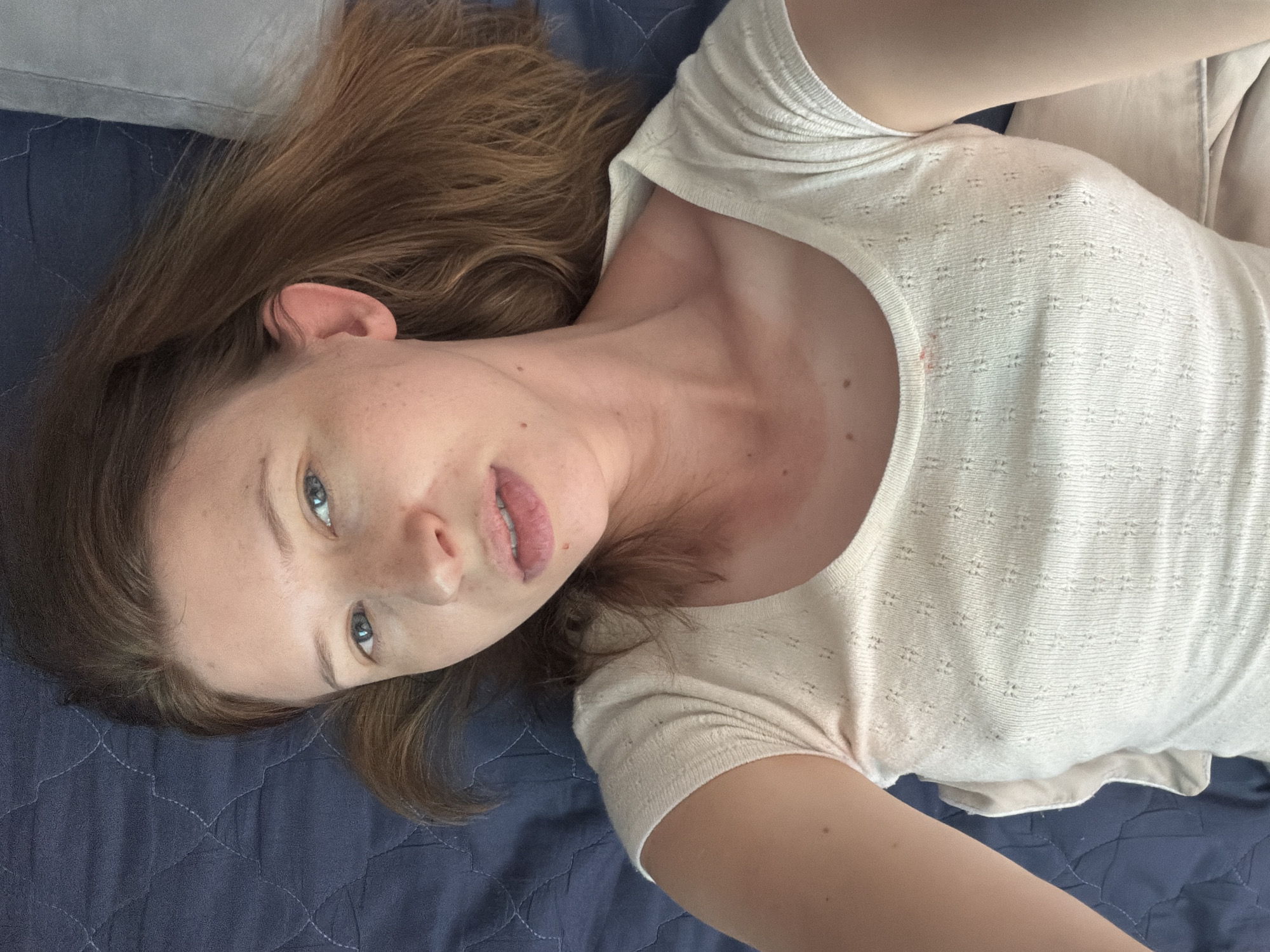 A woman is lying on a dark blue quilted surface, likely a bed or sofa. She is looking directly at the camera with a neutral expression. Her hair is long and a light brown color, spread out around her head. She is wearing a light-colored, short-sleeved top with a textured pattern. The lighting suggests it is daytime, possibly indoors with natural light. There are no other people or objects of significance in the frame. The background is a simple quilted fabric in a dark blue hue. The image appears to be a selfie, given the perspective. The location of Dongducheon-si, South Korea is not visually represented in the image itself, and no elements within the photograph provide any clues to its specific geographical setting.
