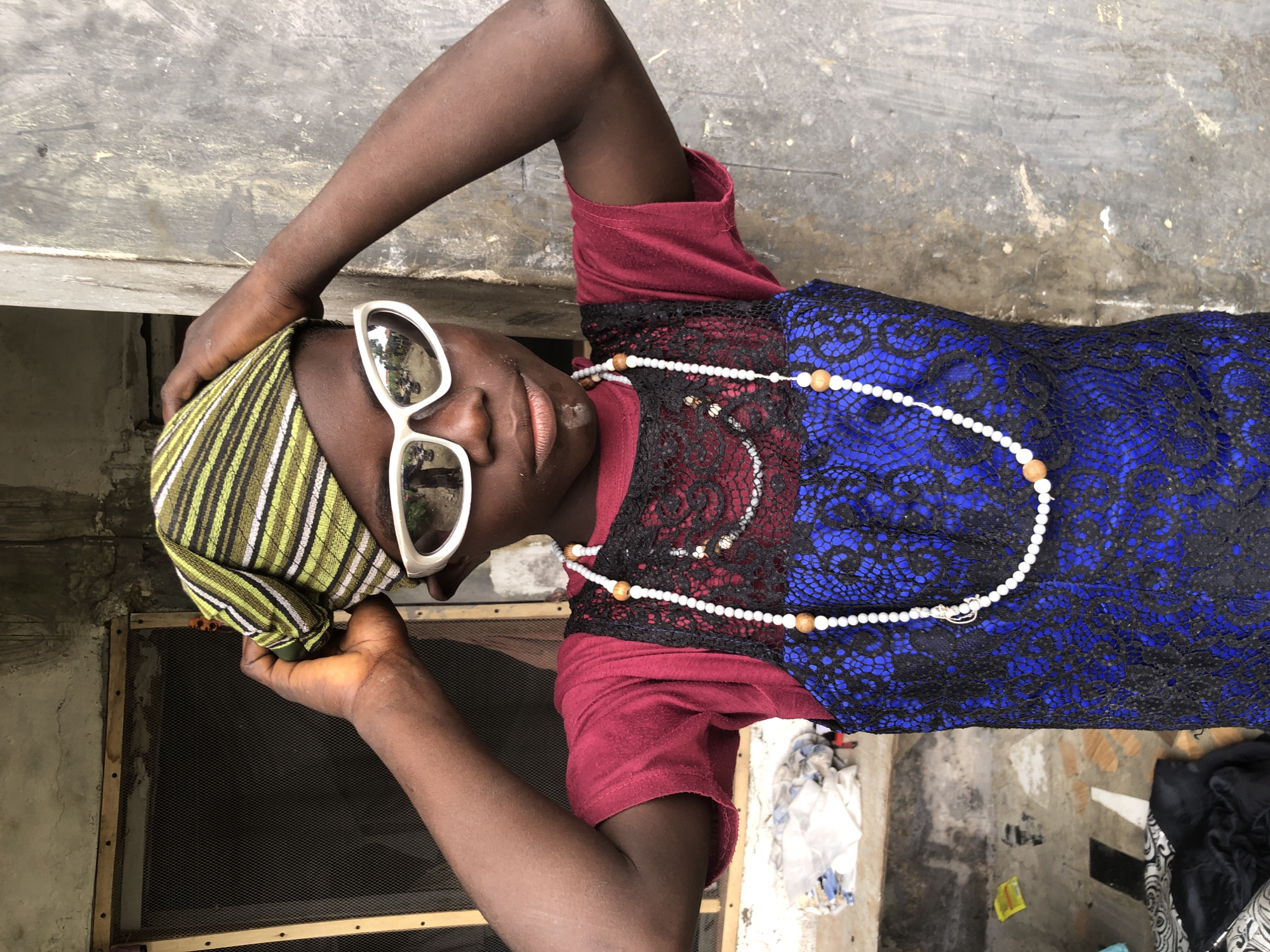 In Ado, Nigeria, a young person stands outdoors, looking at the camera. They are wearing a green and yellow striped headscarf, large white sunglasses, a maroon t-shirt, and a blue dress with a black lace overlay. A white beaded necklace with wooden accents hangs around their neck. Their hands are raised, adjusting the headscarf. The background consists of a weathered concrete wall and a window with a screen. Debris and a pile of fabric are visible on the ground to the left. The lighting suggests it is daytime.