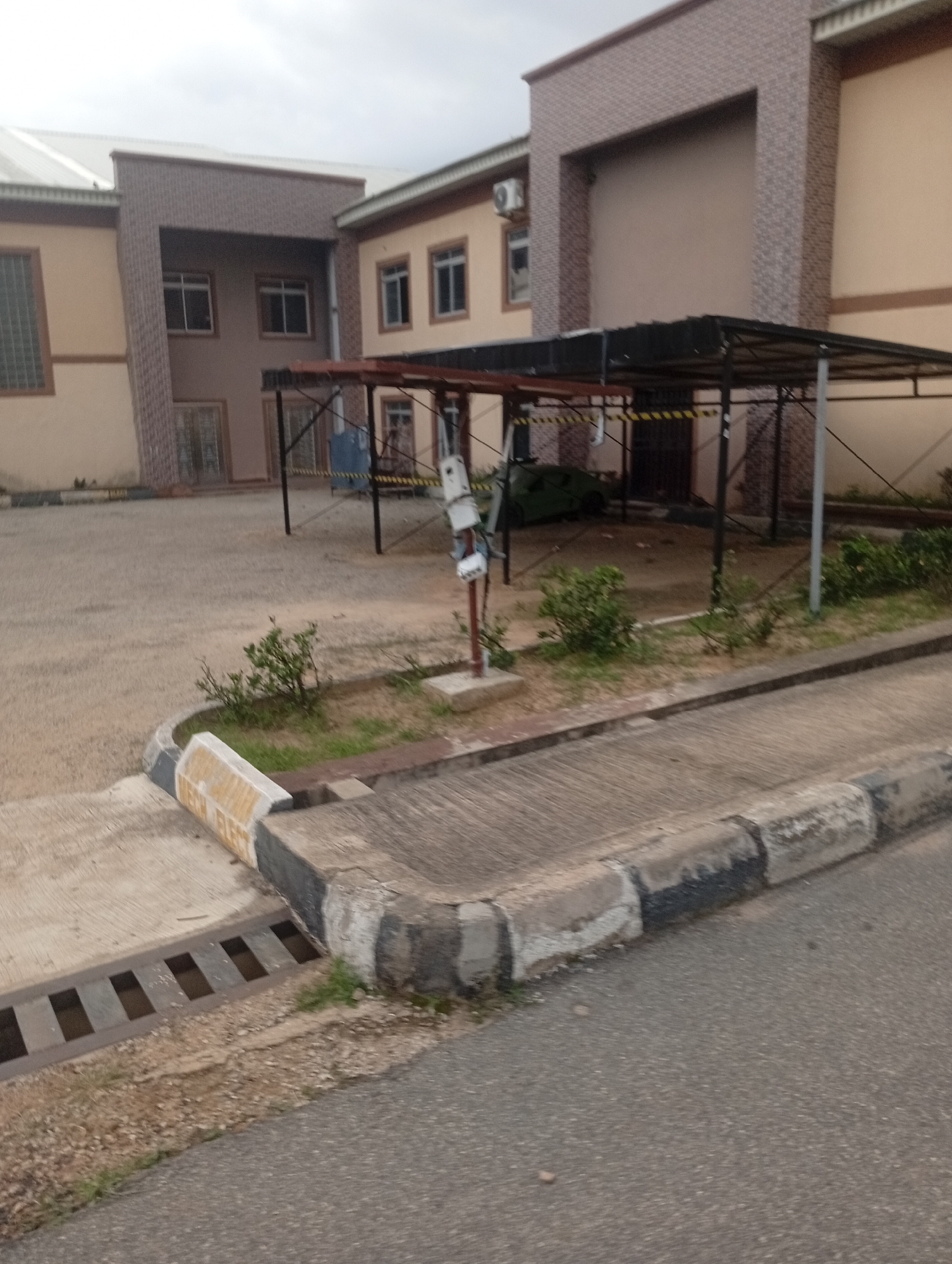 The image depicts a section of a modern building with a light brown facade and a patterned brick accent wall, located in the city of Jos, Nigeria. In the foreground, there is a paved area with a concrete curb and a drainage grate. A covered parking structure with a dark roof and metal supports is visible in the middle ground, with a dark-colored vehicle parked underneath. Several small green bushes are scattered around the paved area. A white pole with some attached equipment stands near the covered parking. The sky is overcast, suggesting a cloudy day. The overall scene appears to be an exterior view of a commercial or institutional building.