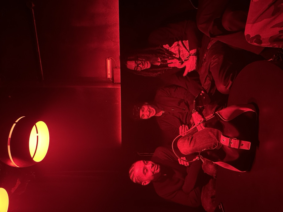 The image shows three individuals in a dimly lit, red-toned room. One person, with dreadlocks and glasses, is making a hand gesture. Another person is wearing a bandana. The third individual has short, light hair. A table is visible with a bag on it. The environment includes wall lights with a circular design.