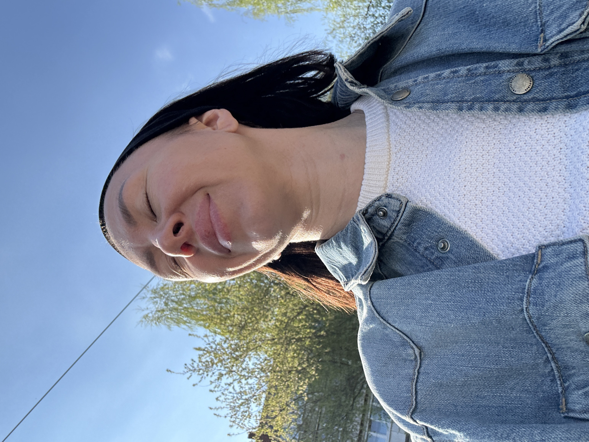 A low-angle, outdoor photo shows a woman with dark hair and closed eyes, smiling slightly, and wearing a white sweater and a denim jacket. Behind her are trees with green leaves against a clear blue sky. The location is potentially in Votkinsk, Russia, but the image doesn't offer specific identifying markers of the city.