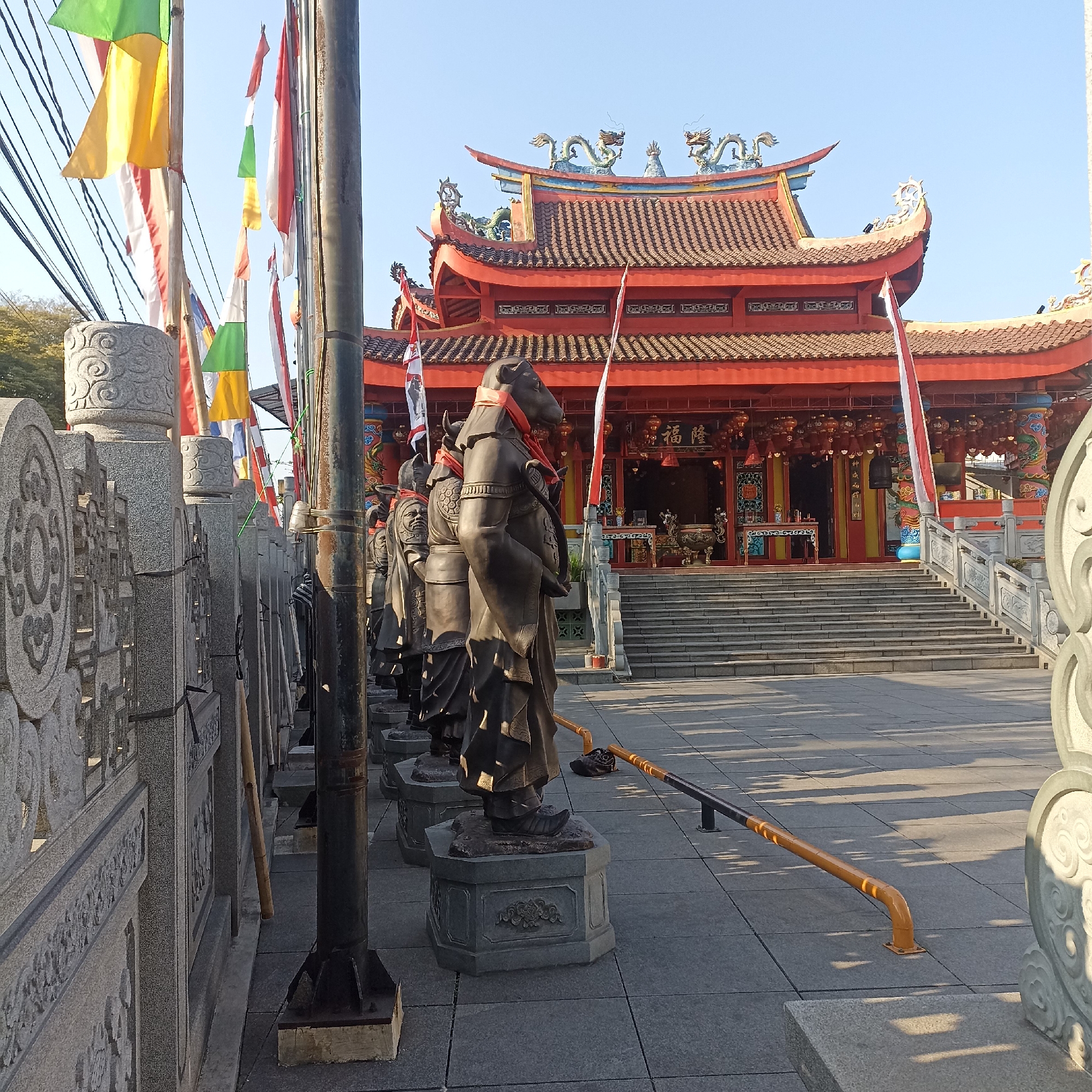 The image depicts the front of a Chinese temple, likely in Magelang, Indonesia, under a clear blue sky. In the foreground, a row of weathered bronze statues of warriors, wearing traditional armor and red sashes, stands on hexagonal pedestals. The statues are arranged in a line, facing the temple entrance. To the left of the statues, a stone fence with intricate carvings runs parallel to the statues. Several Indonesian flags, along with other colored flags, are planted on poles behind the stone fence, fluttering in the breeze. The temple itself is a vibrant red structure with ornate roofs adorned with dragon sculptures. A wide set of stairs leads up to the temple's main entrance, which is decorated with lanterns and traditional Chinese calligraphy. The ground in front of the temple is paved with square tiles, casting long shadows from the statues and a yellow-and-black safety barrier. The scene appears serene and well-maintained, suggesting a place of cultural and religious significance.