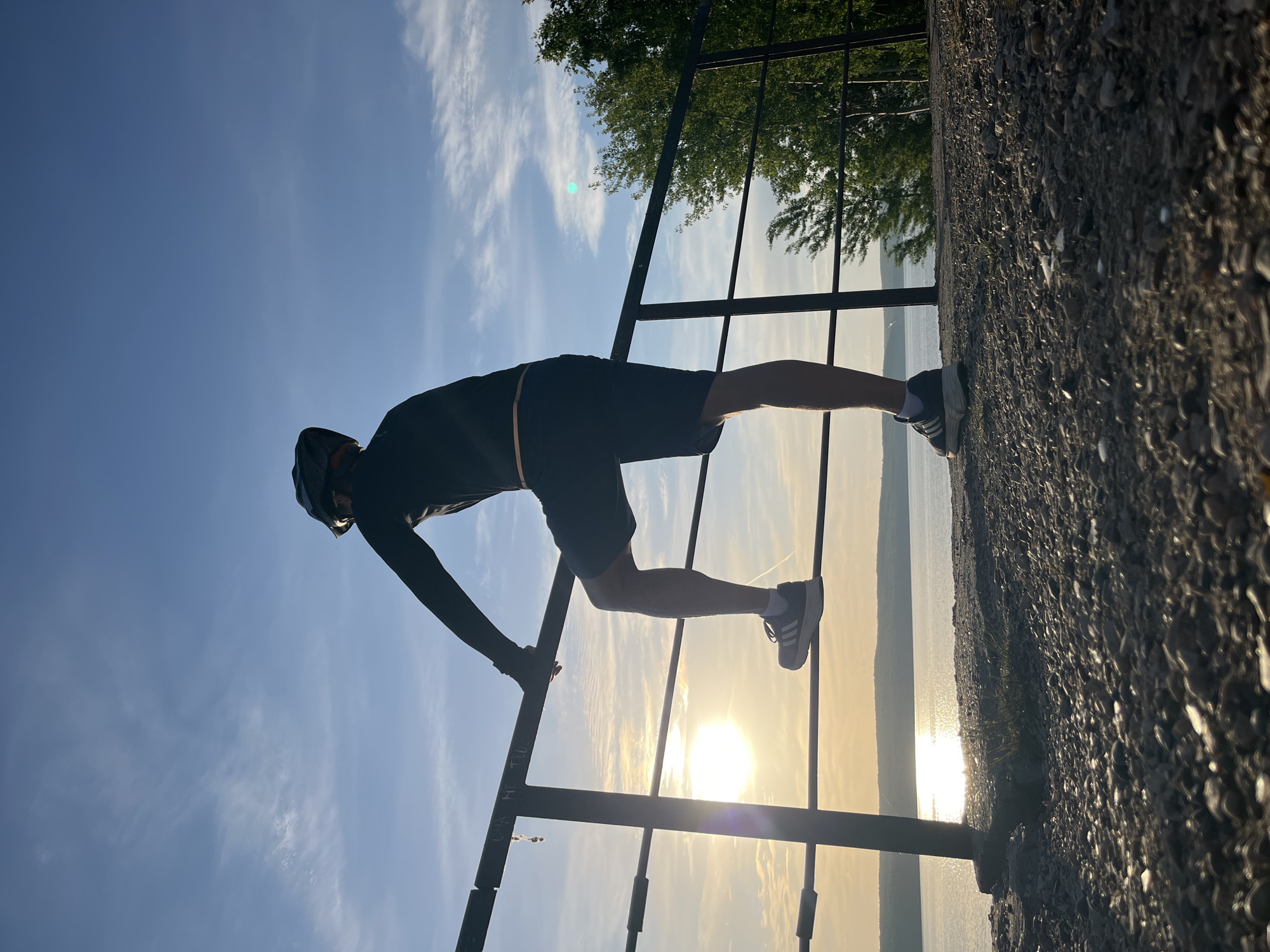 A person is performing a gymnastic maneuver on a railing overlooking a body of water in the city of Chaikovsky, Russia. The individual, silhouetted against the sun, is wearing dark athletic clothing, including shorts, a long-sleeved shirt, and a helmet. Their legs are extended horizontally, with one foot resting on the rail and the other on the ground. The person's body is bent forward, with one arm gripping the railing. The scene is outdoors, with a clear sky and some trees visible in the background. The body of water reflects the sunlight.