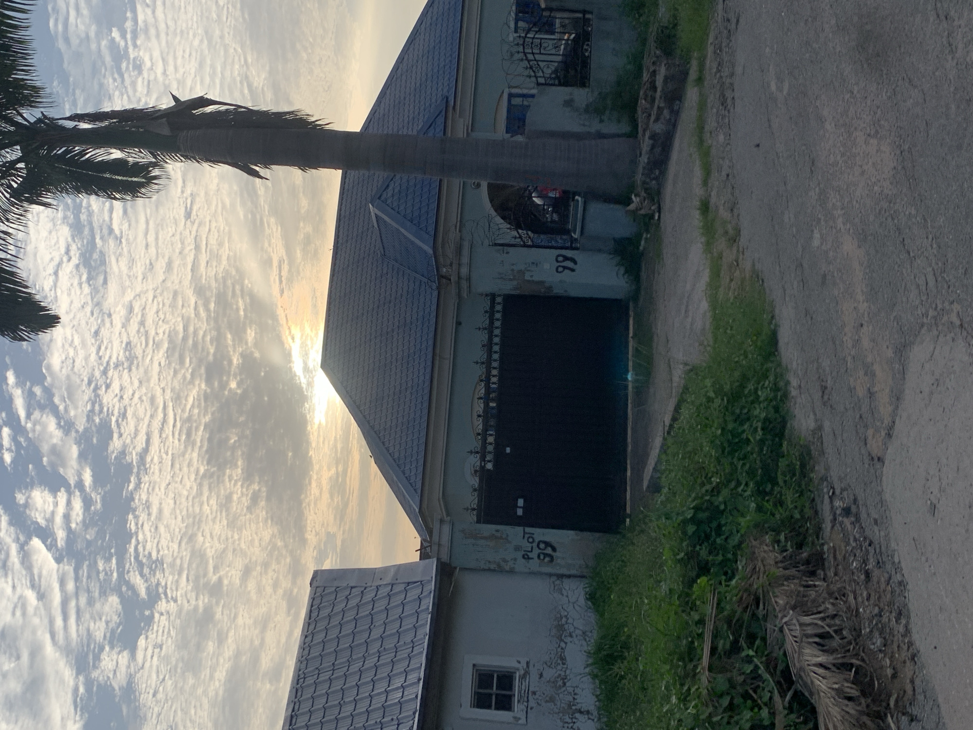 The image shows the exterior of a residential building, likely in Abuja, Nigeria. The building has a metal gate, and above the gate, the number "99" is painted on the wall. A palm tree is located near the building. The sky is cloudy, with the sun's rays visible. A paved road runs alongside the building. Grass and weeds grow on the roadside.