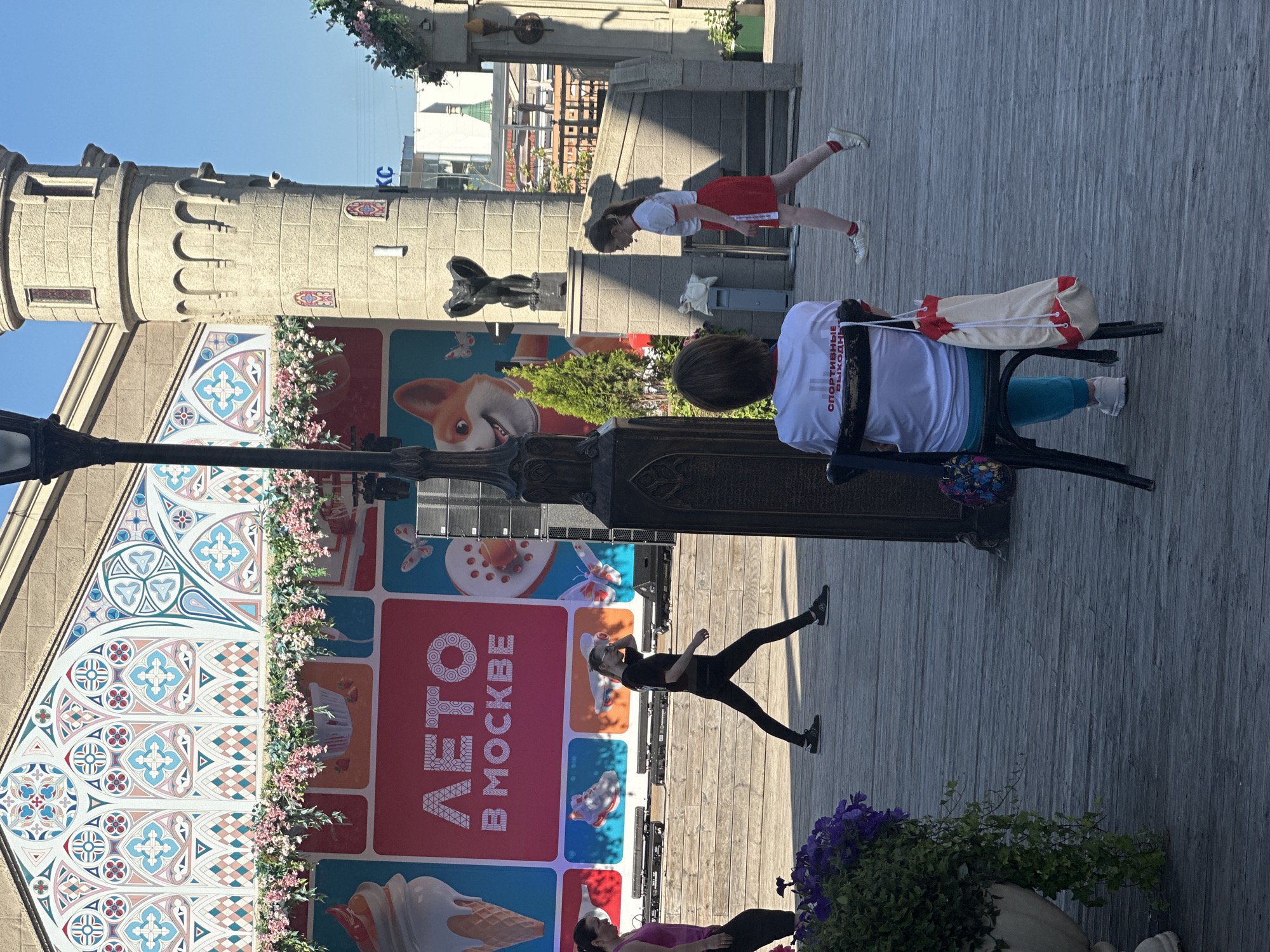 The image depicts an outdoor scene in Moscow, Russia, likely a public space such as a park or a festival area, given the presence of a stage-like structure with a large banner displaying "ЛЕТО В МОСКВЕ" (Summer in Moscow) and images of ice cream, strawberries, and a corgi. The background features a building with architectural elements resembling a castle tower and decorative murals. In the foreground, several people are visible. On the right, a young woman in athletic wear, a white t-shirt with red trim and a red skirt, walks away from the camera. To her left, a woman sits on a dark chair facing away from the camera, wearing a white t-shirt with Russian text that appears to say "СПОРТИВНЫЕ ВЫХОДНЫЕ" (Sports Weekends) and blue sweatpants. She has a light-colored bag with red accents hanging from her chair. In the middle ground, two women are walking on a wooden platform. One woman on the left is wearing a pink tank top and black leggings, while the woman to her right is wearing a black t-shirt and black leggings, engaged in a dynamic pose suggestive of exercise or dance. A tall, dark lamppost is positioned prominently in the center of the frame. Lush green plants and purple flowers are visible in large planters on the left side of the image. The overall atmosphere suggests a sunny day with activities taking place outdoors.