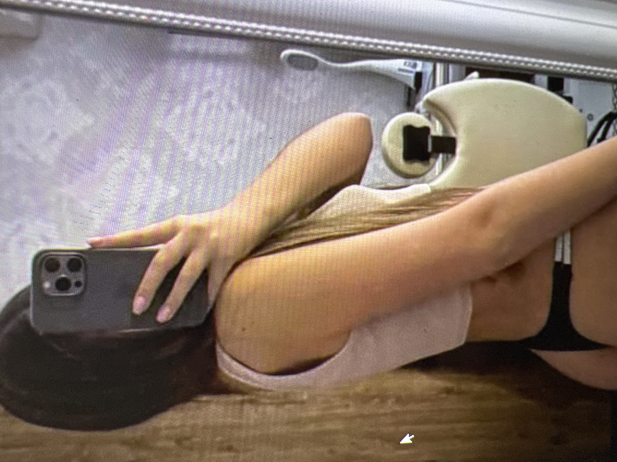 A woman is taking a mirror selfie indoors. She is wearing a light pink top and black bottoms. The background appears to be a room with a wooden textured wall and a large mirror. The mirror reflects a chair with a light-colored cushion and a black frame, and a white electrical appliance. There are no discernible architectural features that would place this image specifically in Castelo Branco, Portugal.