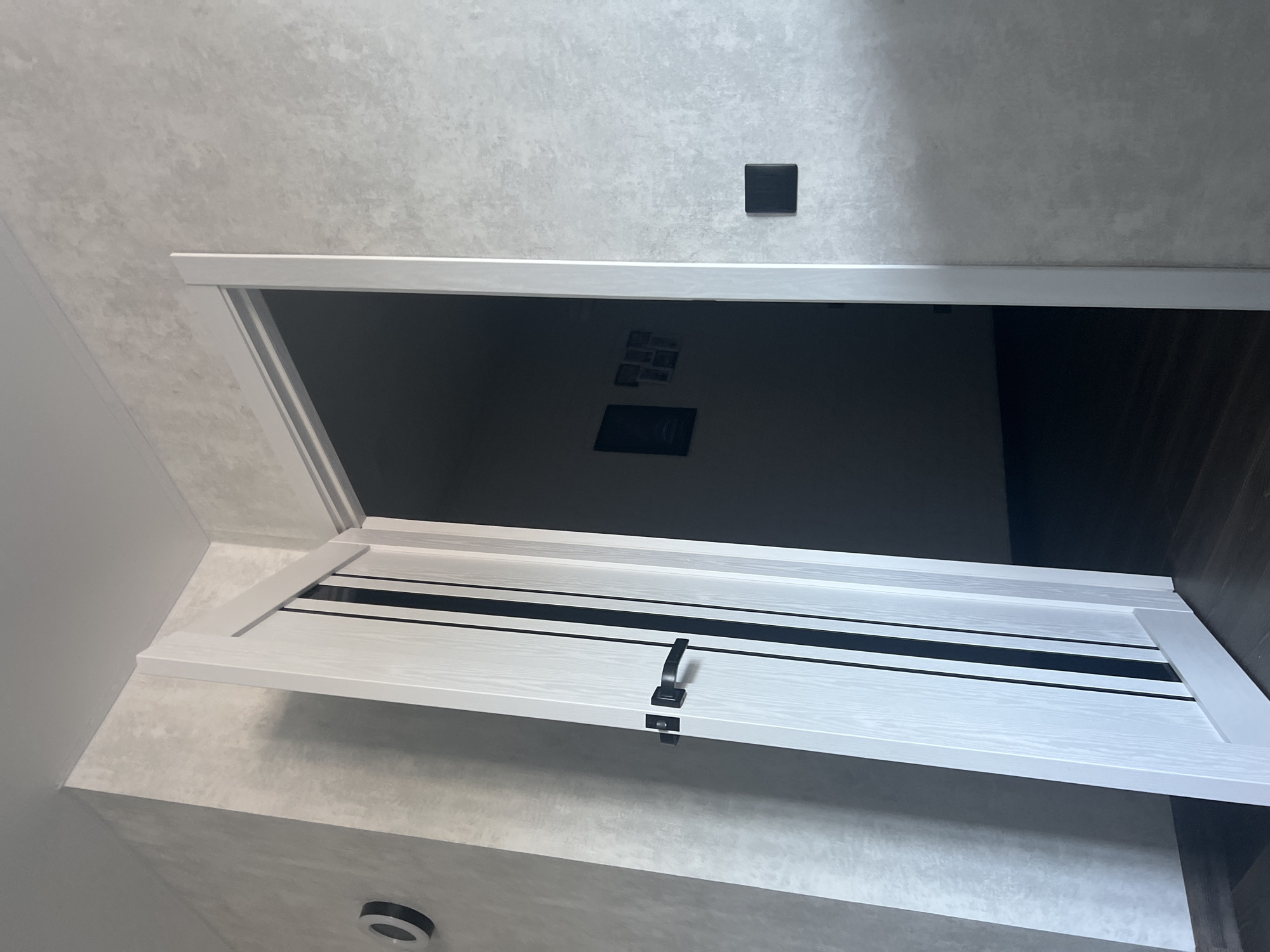 An interior shot, looking towards an open doorway in a modern apartment or hallway. The walls have a textured, light gray finish. To the left, a white door with three vertical black stripes is ajar. The door has a black handle and a black latch mechanism. On the wall to the left of the door, there is a circular, black decorative item with a white ring. To the right of the doorway, a black square electrical outlet is visible on the wall. The floor appears to be dark wood. Through the open doorway, the interior of another room is visible, appearing dark with some framed pictures on the wall. The lighting suggests a somewhat dim environment, possibly from artificial light sources.