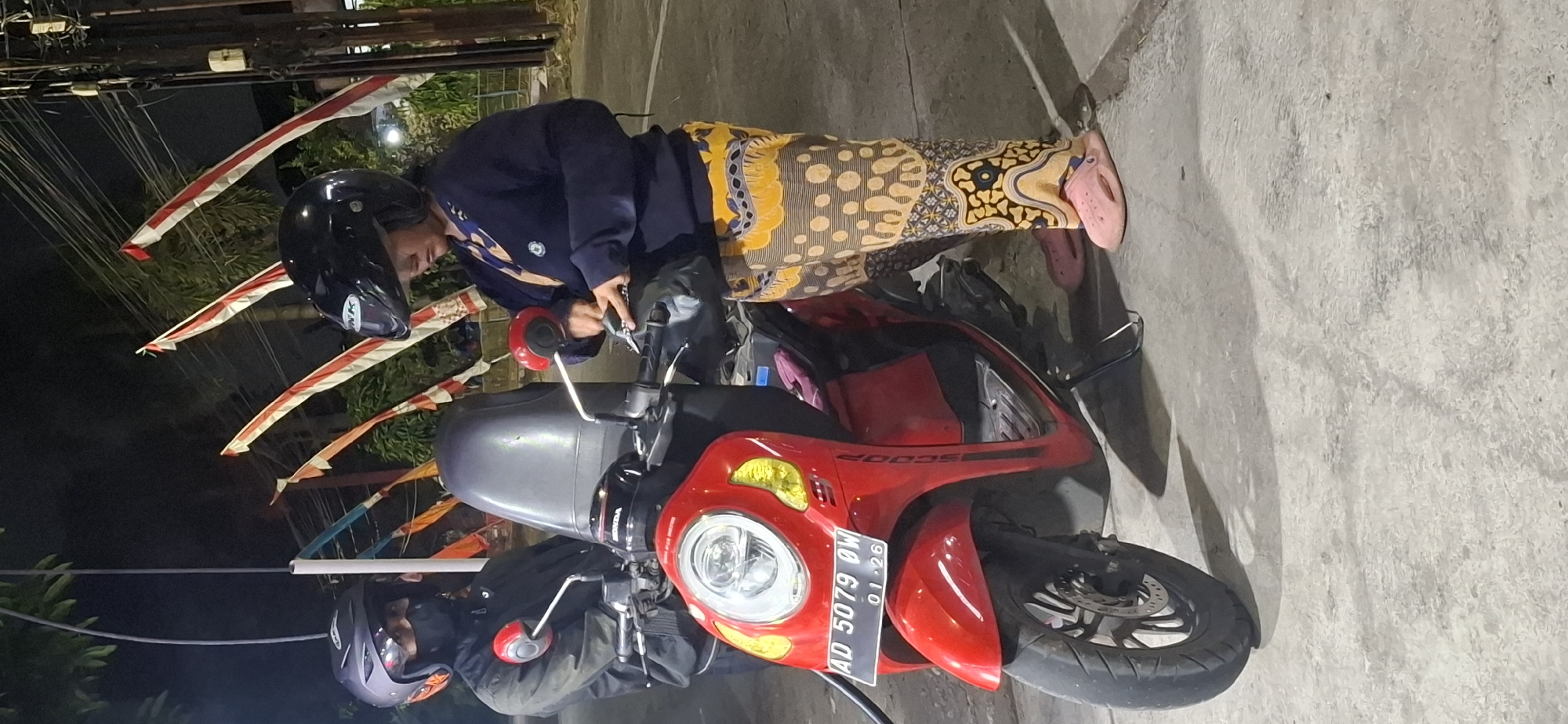 In Desa Gagaksipat, Indonesia, two individuals are standing next to a red scooter. The person on the left is wearing a dark jacket and a black helmet, with a black mask covering their face. They are holding onto the scooter's handlebars. The person on the right is wearing a dark blue jacket and a black helmet. They are wearing patterned trousers in yellow, brown, and white, and pink slip-on shoes. This individual is looking down and appears to be interacting with something held in their hands, possibly a bag or personal item. The scooter has a license plate reading "AD 5079 OW" and "01.26". Red and white flags, likely Indonesian flags, are visible in the background, along with power lines and greenery, suggesting an outdoor, possibly roadside, setting at night. The ground is paved.