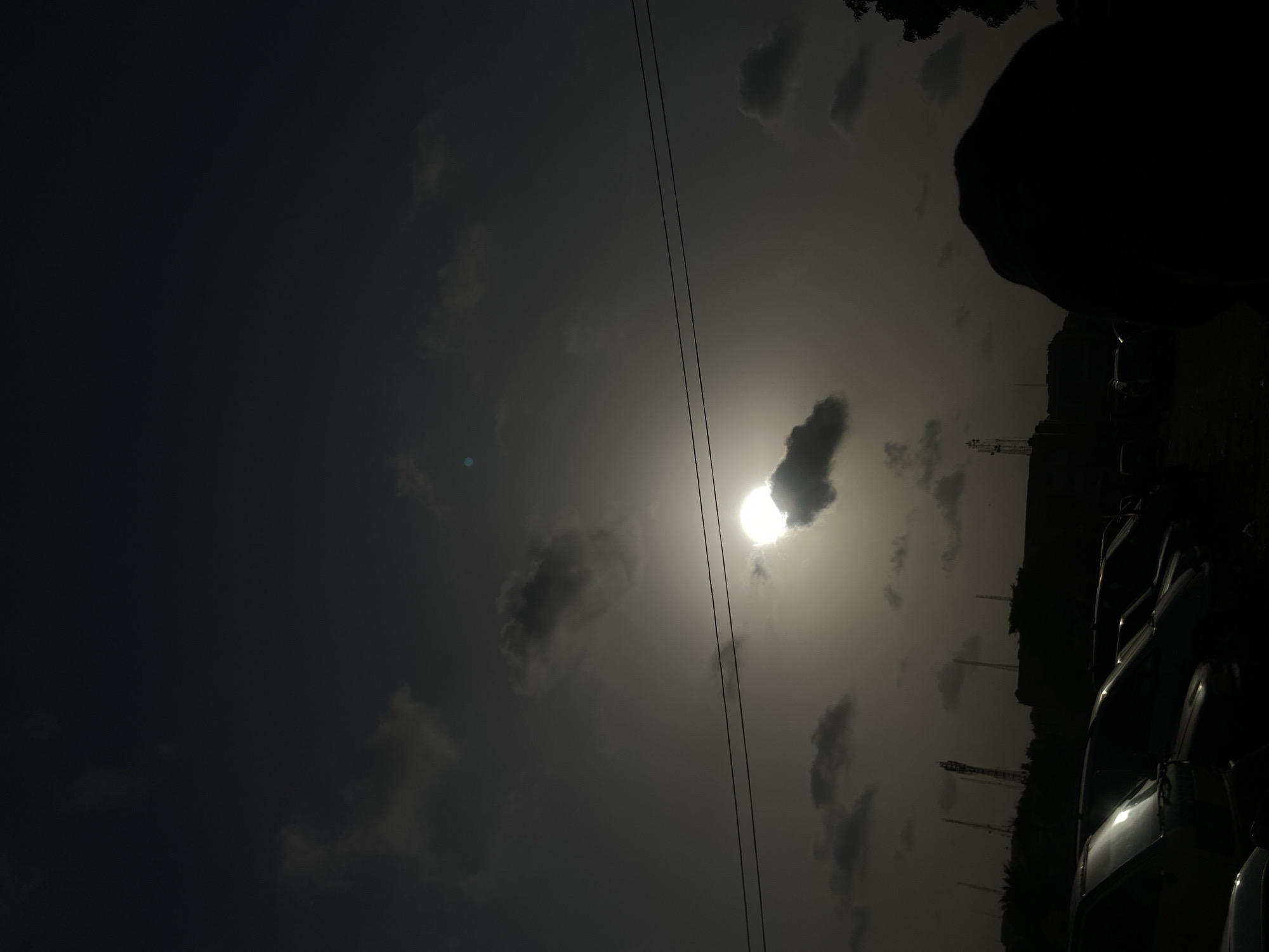 The image shows a dark, possibly overcast sky in Maiburiji, Nigeria. The sun is partially obscured by clouds. Power lines run across the upper center of the frame. Silhouetted structures, including what appear to be communication towers, are visible on the right side. The bottom right portion of the image shows the outlines of vehicles in a dark environment. No people are visible.