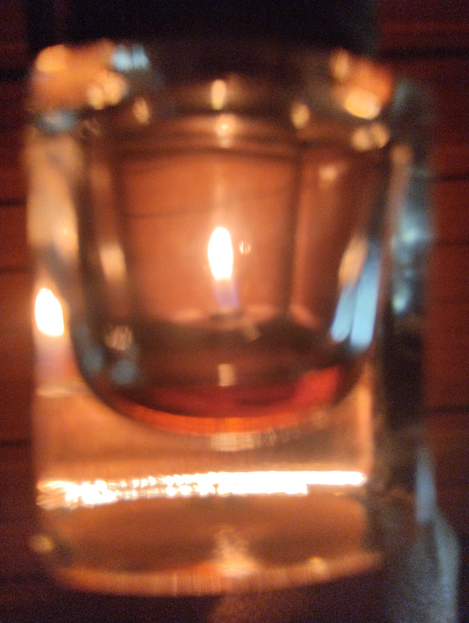 This media file depicts a close-up, blurry shot of a single lit candle in a glass votive holder. The candle flame is visible and emits a warm glow. The votive holder itself appears to be made of clear glass and is filled with a reddish liquid, likely melted wax or oil. The background is dark and indistinct, suggesting an indoor setting at night. Hints of wooden textures can be seen in the background, possibly indicating the candle is placed on a wooden surface or near wooden furniture. The overall mood is calm and intimate, likely captured during a quiet evening or as part of a decorative ambiance. There is no visible text within the image. The location context of Urkovo, Russia, does not provide specific visual cues within the image itself.