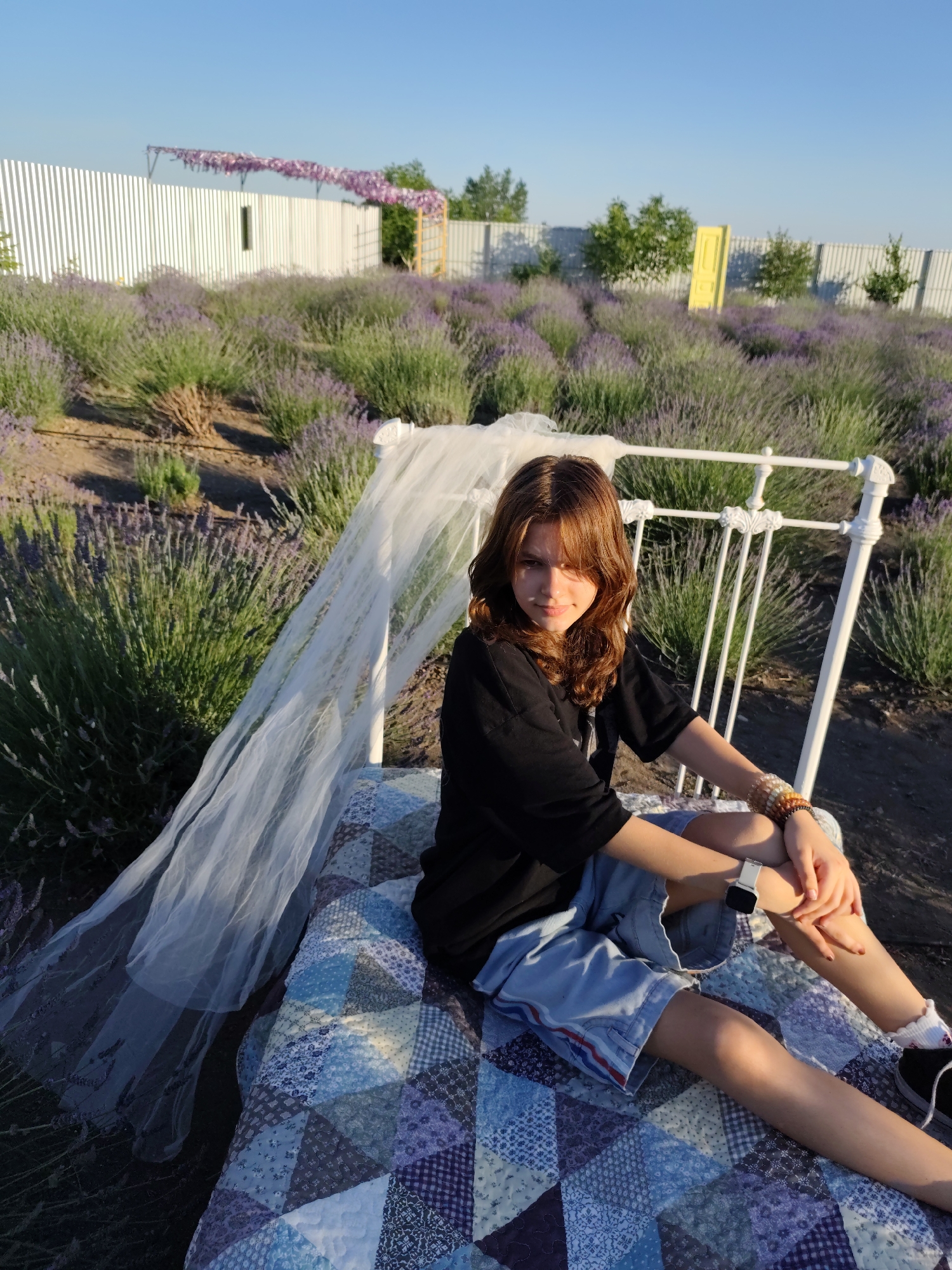 A young woman with shoulder-length brown hair and bangs is sitting on a quilted blanket spread over a bed frame in a lavender field. She is wearing a black t-shirt and light blue shorts, and a white watch on her left wrist. She has bracelets on her right wrist. A sheer white veil drapes over the bed frame and extends to her left. The background is a vast field of purple lavender under a clear blue sky. A white corrugated metal fence runs along the horizon, with trees and a yellow door visible in the distance. The sun is low in the sky, casting a warm glow and long shadows, suggesting it is late afternoon or early evening. The overall mood is serene and peaceful. There is no visible text in the image.