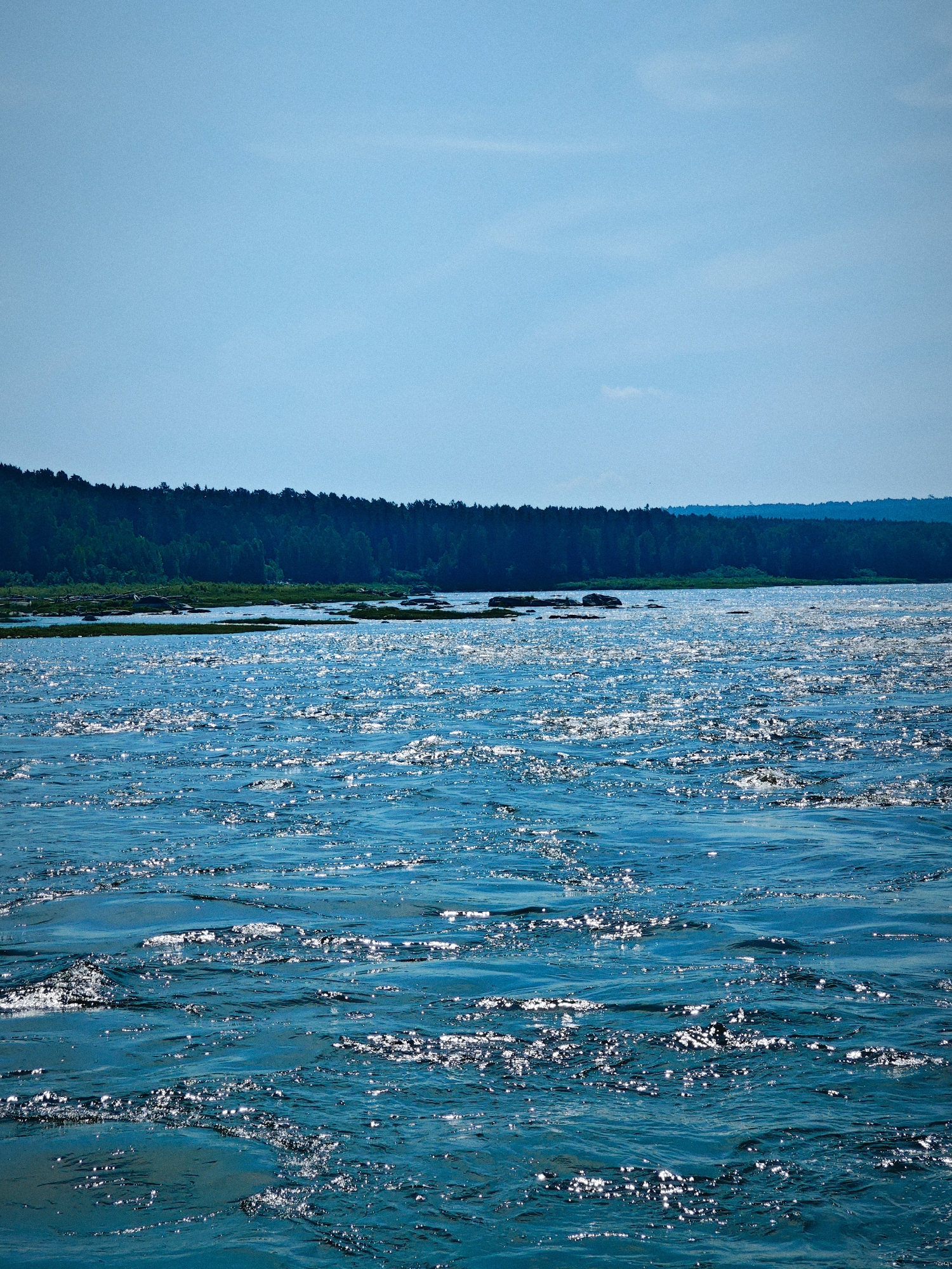 The image depicts a wide expanse of choppy, blue water, reflecting the bright sunlight with numerous glittering highlights. In the background, a dense, dark green forest stretches horizontally across the horizon, creating a stark contrast with the bright water and sky. The sky is a clear, pale blue with a few faint, wispy clouds visible in the upper portion. The scene is likely a natural landscape, possibly a river or lake in the city of Porog, Russia, given the hint provided. There are no people or specific objects clearly identifiable in the image, other than the natural elements of water, forest, and sky. The overall impression is one of a sunlit, natural environment.