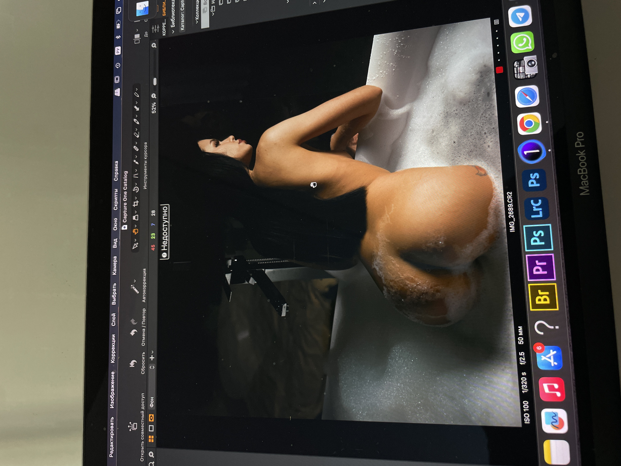A photograph of a MacBook Pro screen displays an image of a nude female torso in a bathtub filled with foam. The photograph is open in a photo editing program, Capture One Catalog. The image is dark, with the subject's skin tones appearing warm. The top right corner displays app icons. The operating system appears to be macOS. The computer is on a white surface. There are no specific elements indicating that this image or scene originated in Kazan, Russia.