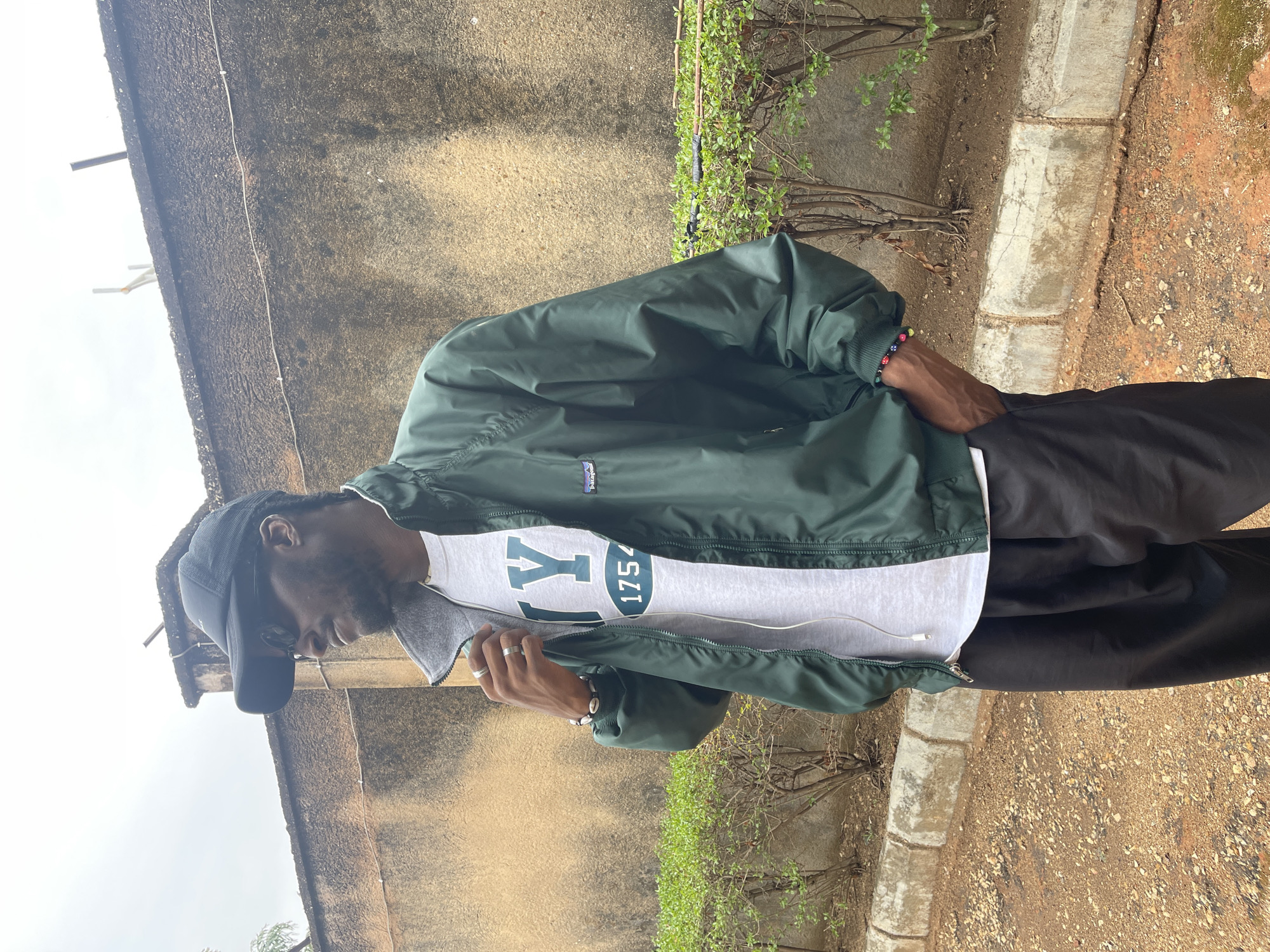 A person stands outdoors, against a backdrop of a weathered wall and sparse greenery, possibly in the city of Jos, Nigeria. The individual is wearing a dark baseball cap, a green jacket over a grey t-shirt with "Y 1754" printed on it, and dark pants. They appear to be looking down, perhaps occupied with something in their hands. The setting includes a paved edge adjacent to the wall, indicating a possible sidewalk or walkway, with an unpaved surface nearby, suggesting a garden or undeveloped area.