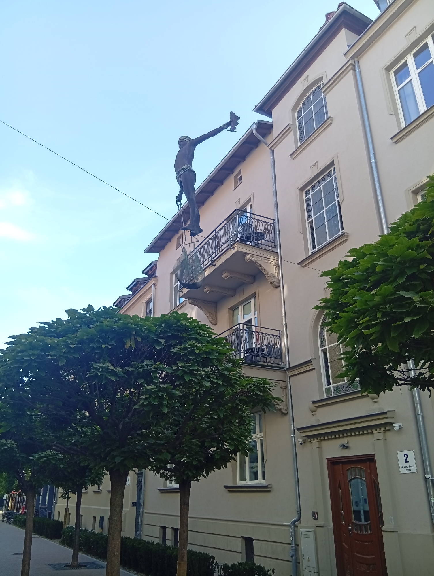 The image depicts a bronze sculpture of a man suspended by ropes from a building's gutter in Sopot, Poland. The sculpture appears to be a window cleaner, with one arm extended as if reaching for something. The building is a light-colored, multi-story structure with arched windows and balconies. Several trees with green foliage are visible in the foreground, lining a sidewalk. A street number "2" is visible on the building near the entrance. The sky is clear and blue.