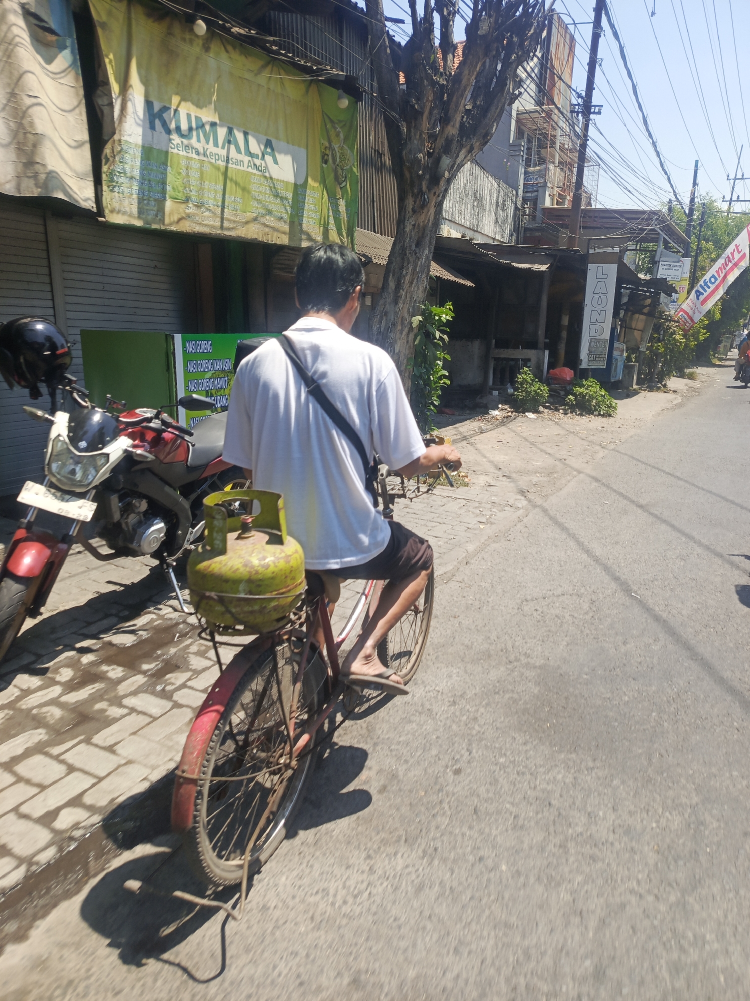 A person is riding a bicycle with a green gas cylinder strapped to the back in the city of Wonocolo, Indonesia. The person is wearing a white t-shirt and brown shorts. The bicycle is red and has a spoke wheel. The gas cylinder is yellow-green. To the left of the bicycle is a red and white motorcycle. The background shows a building with a banner that says "KUMALA Selera Kepuasan Anda". The road is paved and there are trees and power lines in the background. The sun is shining and the sky is blue.