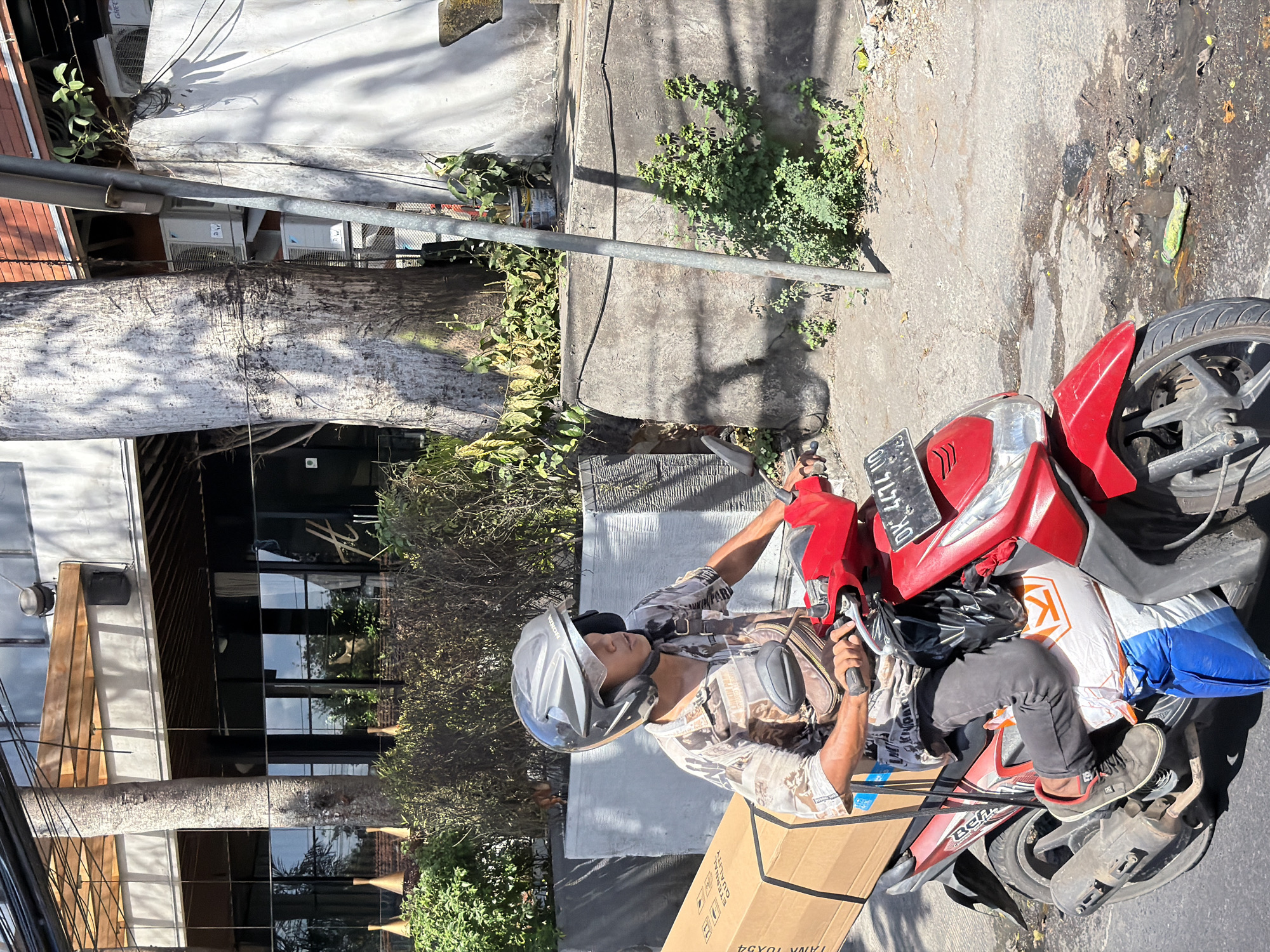In Kuta Selatan, Indonesia, a person wearing a helmet rides a red scooter down a street. They are carrying a large cardboard box secured to the side of the scooter. The scooter also has bags and a white and orange package attached to it. The person is wearing a patterned shirt, dark pants, and gray sneakers. The street is paved with asphalt and shows signs of wear. In the background, there are buildings with large windows, trees, and vegetation. The scene is illuminated by bright sunlight, casting shadows on the ground.
