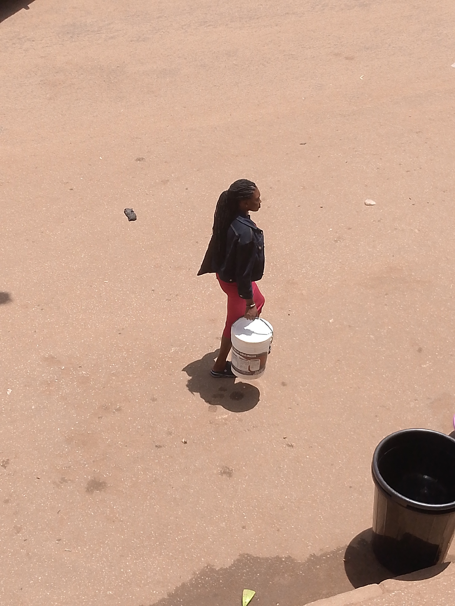 A medium-shot aerial view depicts a woman walking on a concrete surface in Bukuru, Nigeria. The woman has long braided hair and is wearing a dark jacket and red pants, carrying a white bucket with apparent product labeling in her right hand. The environment includes a piece of debris or litter and a black plastic bucket placed in the lower right corner of the shot.