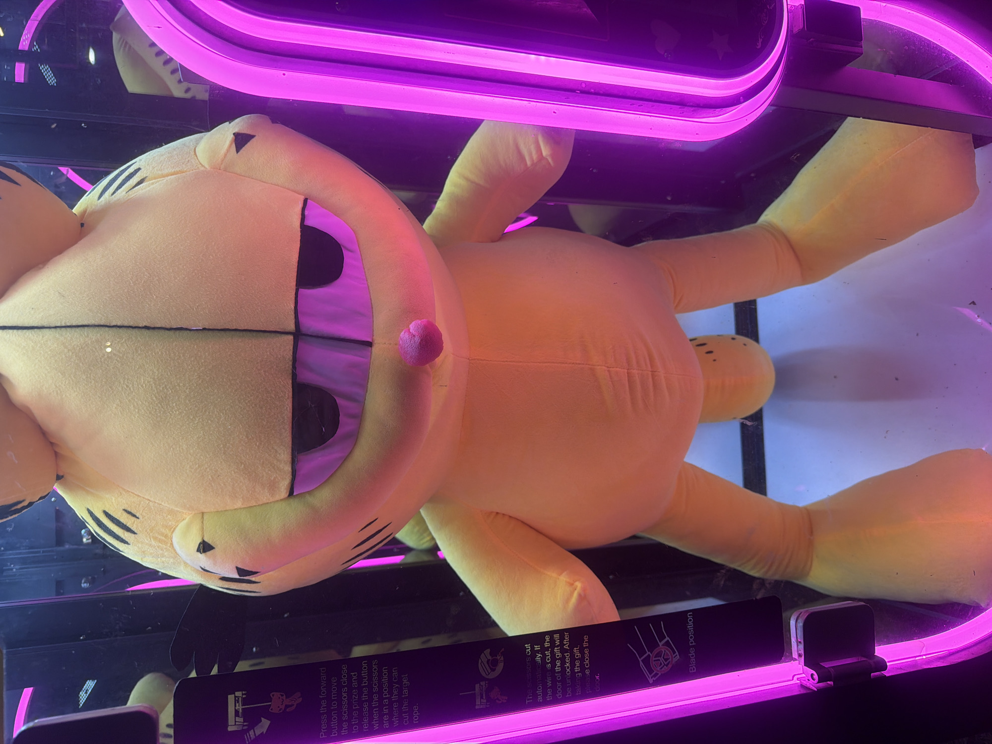 A large yellow and orange Garfield plush toy is prominently displayed inside what appears to be an arcade prize game. The plush is lying on its back, taking up a significant portion of the game's enclosure. Its iconic striped design and cheerful expression with a pink nose are clearly visible. The interior of the game machine is brightly lit by intense purple neon lights, casting a vibrant glow over the plush and reflecting off the clear glass panels.
The game is a "cut the rope" style machine, with instructions visible on a black panel at the bottom of the enclosure. The text describes how to play: "Press the forward button to move the scissors close to the prize and release the button when the scissors are in a position where they can cut the target rope." It further explains the mechanics: "The scissors cut automatically. If the wire is cut, the door of the gift will be unlocked. After taking the gift, please close the door." Another label indicates "Blade position." The overall setting suggests an entertainment venue like an arcade or a family fun center, given the nature of the prize and the game itself. The bright, artificial lighting suggests an indoor, possibly evening, environment.