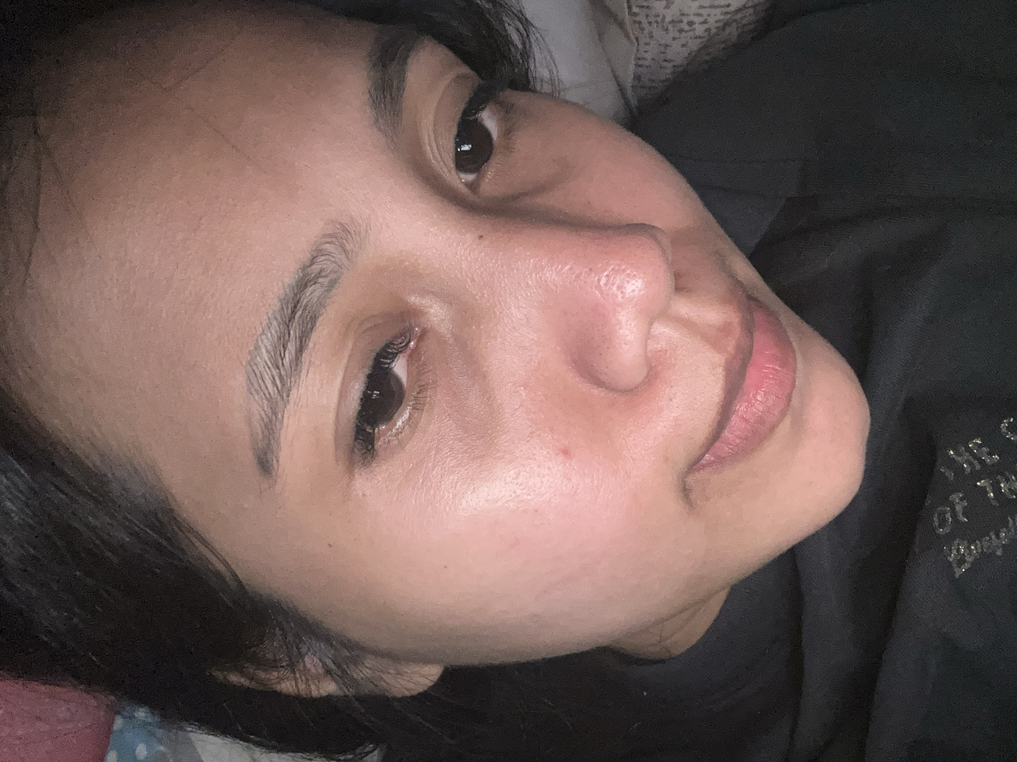 The image is a close-up, upward-facing shot of a young woman's face. She has dark hair framing her face and is lying down. Her eyes are open, with one looking directly at the camera and the other slightly averted. Her eyelashes are long and dark. She has natural eyebrows and a visible mole on her cheek. Her nose is slightly pink, and her lips are parted, revealing a soft pink hue.
The lighting suggests it is likely nighttime or indoors. The background is dark, with a hint of patterned fabric visible in the upper left corner and a black garment covering her shoulder and chest. The garment has some subtle, shimmering text on it, which appears to be part of a design or logo.
The overall impression is intimate and personal, with the subject looking relaxed and possibly contemplative. There are no strong indications of a specific activity or event, other than the act of being photographed. The location context of Depok, Indonesia, does not provide specific visual cues within the image itself.