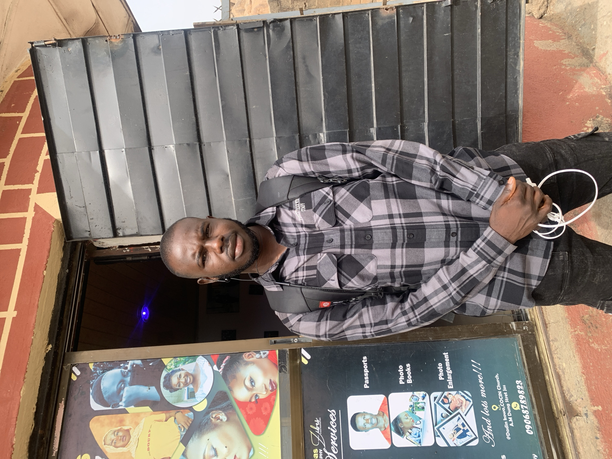 A man wearing a plaid shirt stands in front of a photography studio in Jos, Nigeria. He is positioned in the doorway of "Fibas Services," a photography studio that offers passports, photo books, and photo enlargement. The studio's sign advertises its location as "@Dadin kowa, COCIN Church, A.M Dung Street Jos" and provides the phone number 09068789823. The man is holding a white cord in his hand. He wears a backpack. The studio entrance is framed by a black metal door with horizontal slats. The building's exterior is a mix of brick and painted surfaces. A blue light is visible inside the studio.