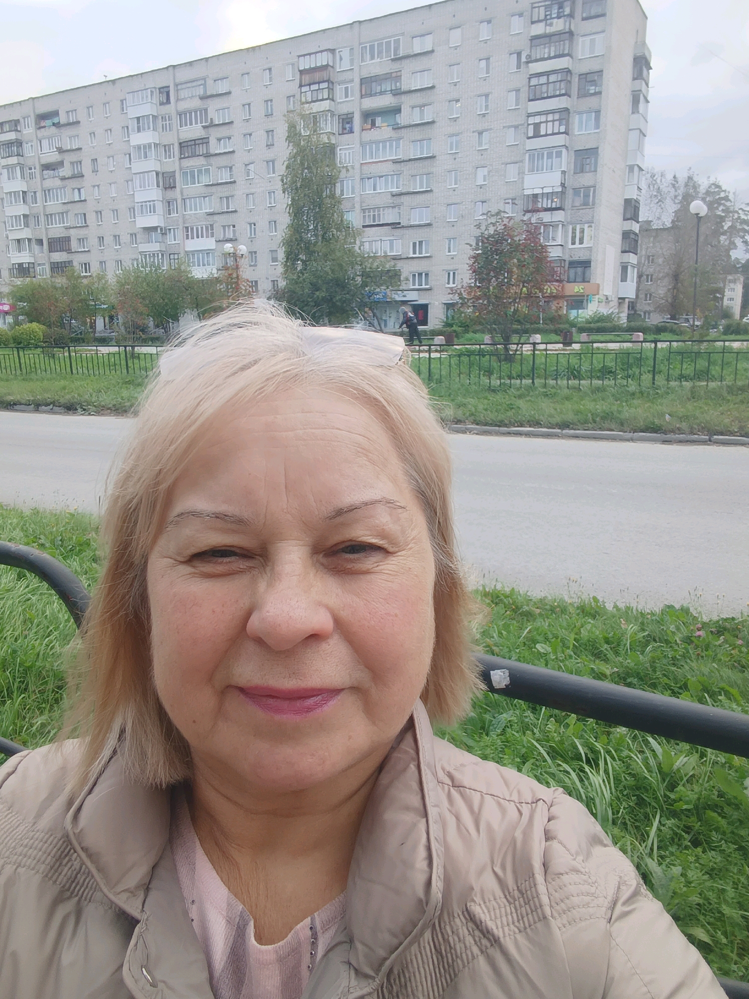 A middle-aged woman with blonde hair and fair skin is taking a selfie outdoors. She is wearing a light brown quilted jacket over a pink shirt. Her lips are painted with a berry-colored lipstick. Behind her, a paved road is visible, bordered by a green grassy area with a black metal fence. In the background, a tall, gray apartment building with many windows dominates the scene. Trees with sparse autumn foliage are also present. The sky is overcast. The location is the city of Asbest, Russia, specifically near the apartment building on Pionerskaya Street, as indicated by identifying features of the building and its surroundings that match existing imagery of that location.