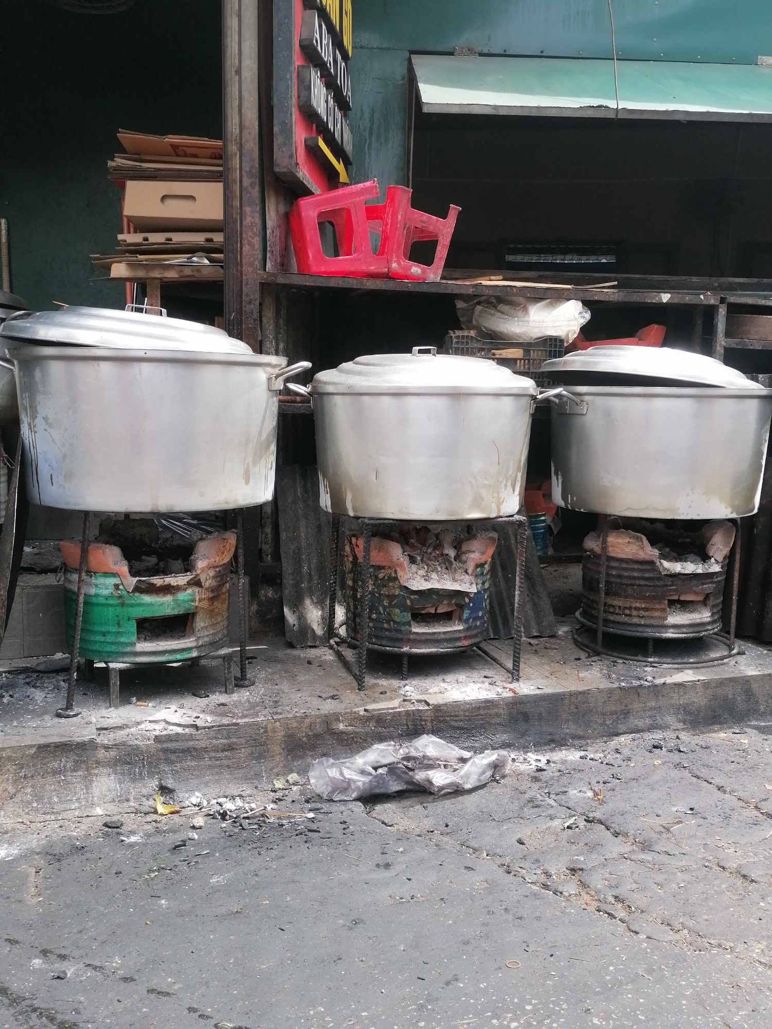 The image shows an outdoor scene in Da Lat, Vietnam, featuring a row of three large metal pots on individual charcoal stoves. The pots appear to be used for cooking, likely food. The stoves are rustic, made from what seem to be repurposed metal drums lined with bricks, and show signs of heavy use with ash and debris around them.
In the background, there is a covered area that could be a shop or food stall. Shelves are visible, stacked with cardboard boxes and other items, including a red plastic stool. A sign with Vietnamese text is mounted above the stall area. The ground in front of the stoves is paved and appears dirty, with scattered debris, ash, and some plastic waste.
The overall impression is of a functional, perhaps modest, food preparation area on a city street, indicative of local street food culture. There are no people visible in the frame.