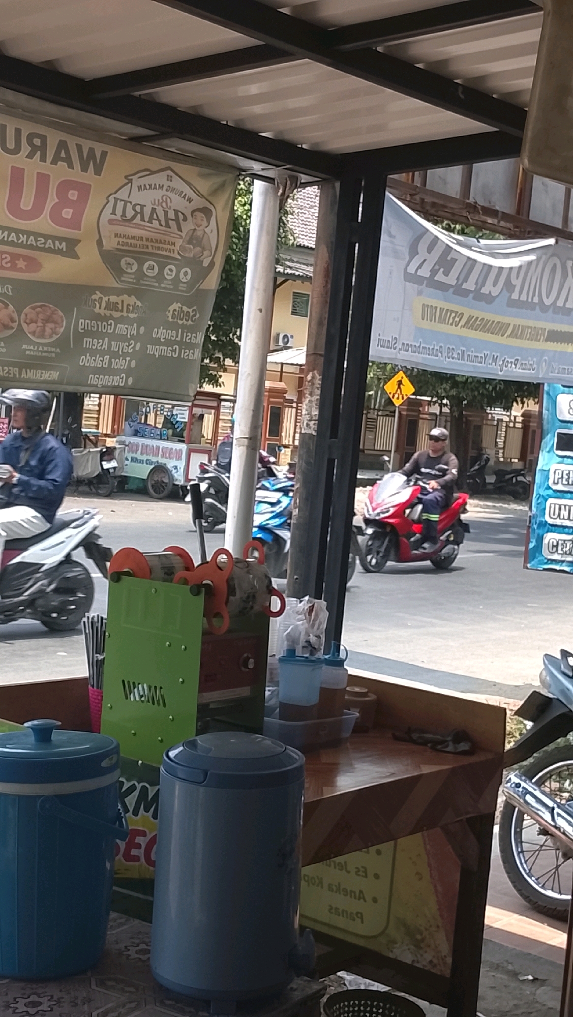 The image depicts a street scene in Slawi, Indonesia, likely near a food stall. In the foreground, there are two large blue coolers and a wooden table with various containers and items. Behind the table, a green and red juicer machine is visible, along with stacks of straws. A street with several motorcycles and a small cart selling drinks is in the background. A red motorcycle with a rider is prominently displayed in the middle ground, and another rider on a white motorcycle is seen in the left of the frame. Two banners are hanging overhead, one featuring a cartoon of a chef and the other with text that is difficult to read. The overall impression is of a busy street setting with food and drink vendors.