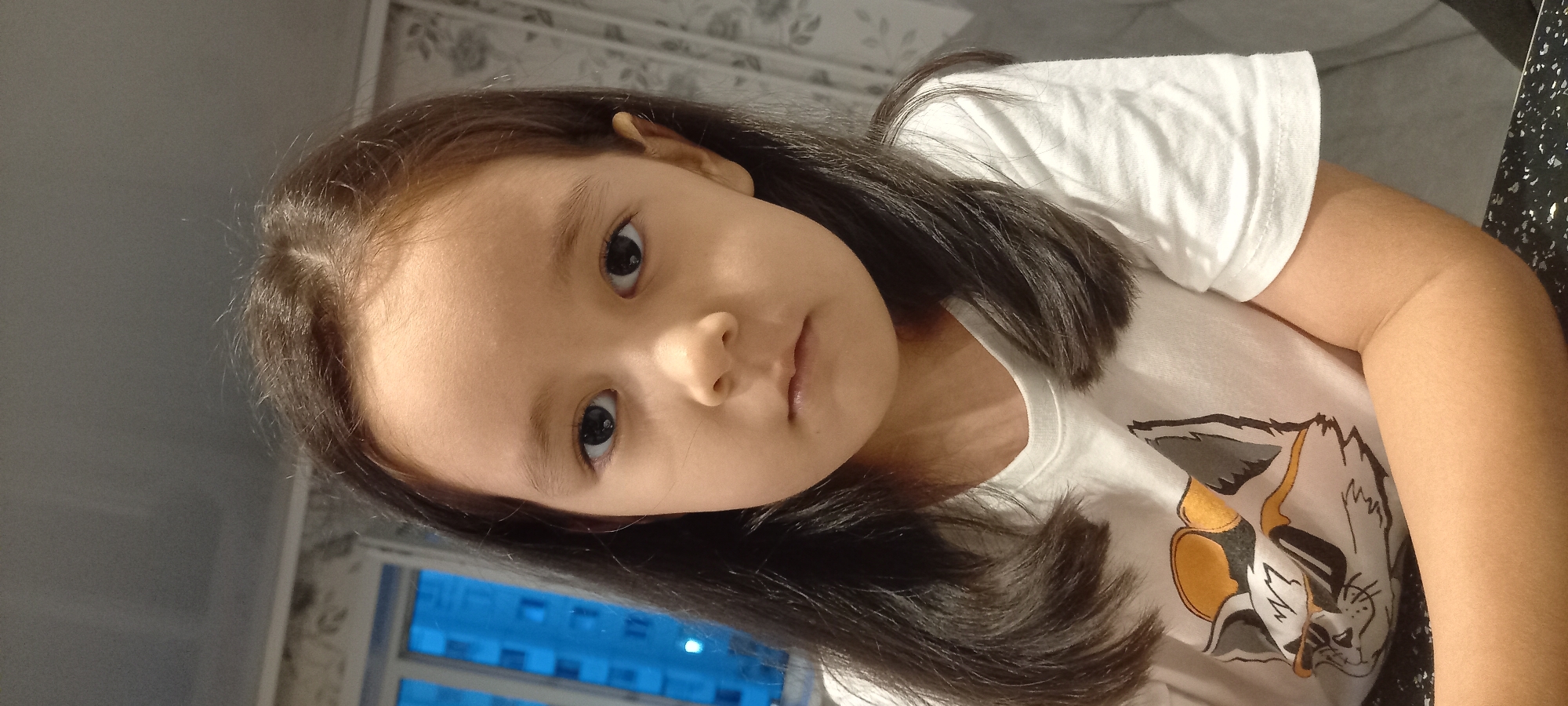 The image features a close-up, vertically oriented shot of a young girl. She has dark hair and is looking directly at the camera with a neutral expression. She is wearing a white t-shirt with a cartoon character graphic. The background is softly blurred, showing what appears to be an indoor setting with patterned wallpaper and a window with blue light filtering through, suggesting it might be evening or nighttime. The lighting in the image is soft, with some shadows cast on the girl's face and hair, possibly from natural light coming from the side. There is no visible text in the image. The overall impression is of a casual, indoor portrait.