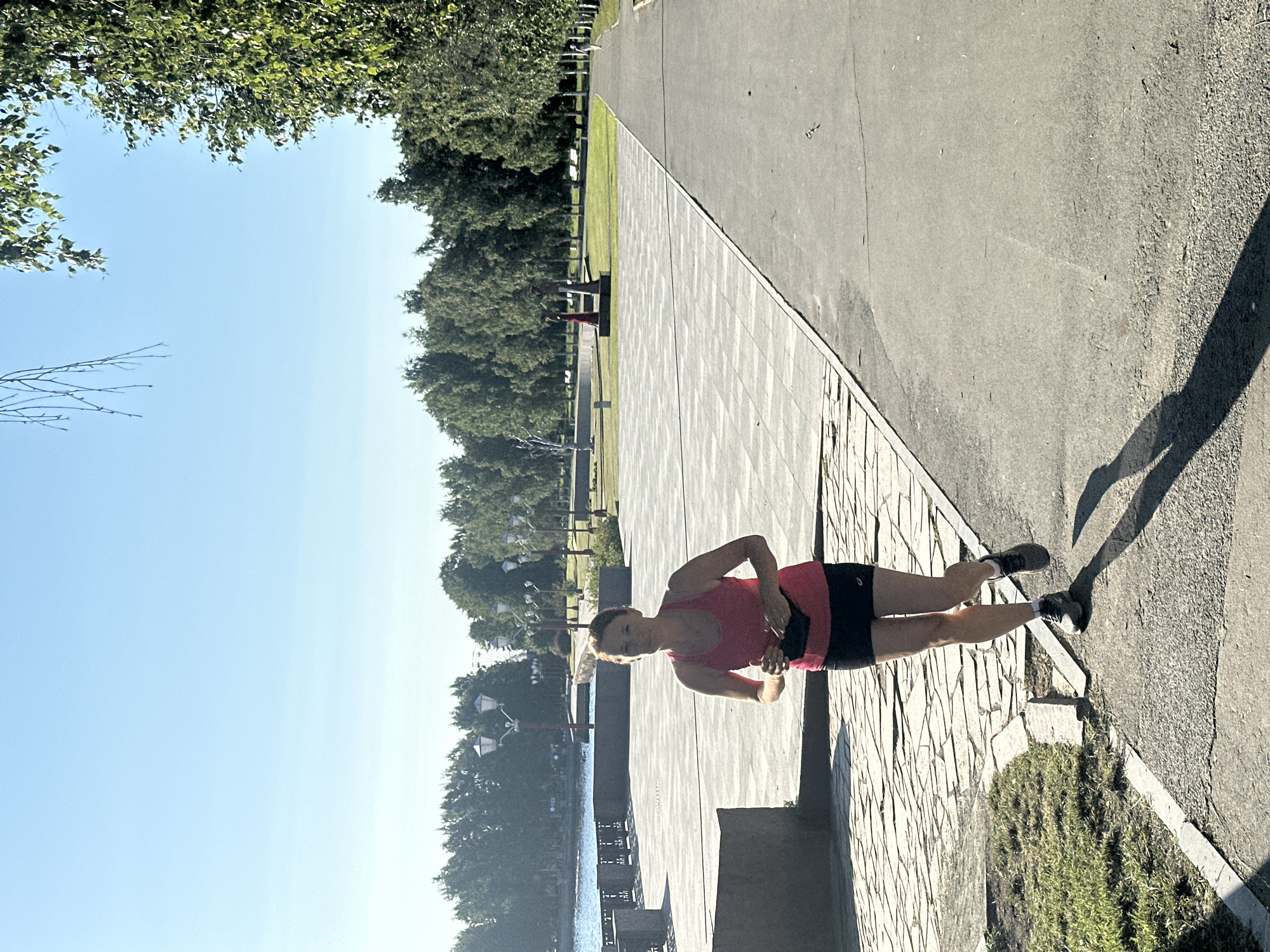 A woman in a pink tank top and black shorts runs on a paved path next to a park in Petrozavodsk, Russia. The path is bordered by a stone walkway and grass on one side and a large paved square on the other. In the background, trees line the edge of a body of water, and streetlights dot the park. The sky is clear and blue. The woman is casting a long shadow on the path.