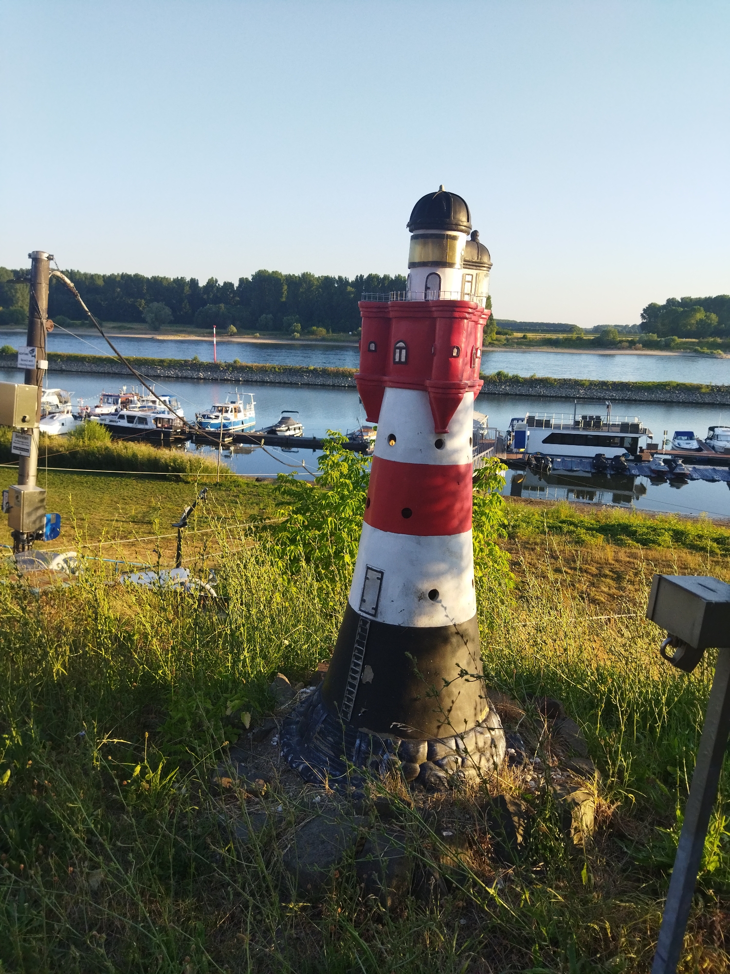 An eye-level, medium shot captures a decorative lighthouse situated on a grassy embankment overlooking a calm river in Leverkusen, Germany. The lighthouse, painted in alternating red and white horizontal bands with a black base and top, features a decorative ladder on its side. Behind it, the river is dotted with various boats, including motorboats and larger houseboats docked at the water's edge. The opposite bank of the river is lined with a dense forest, and the sky above is a clear, pale blue. To the left of the lighthouse, a metal pole with electrical boxes and wires is visible, indicating infrastructure near the riverfront. The foreground is dominated by tall, dry grass illuminated by the warm glow of the setting sun.