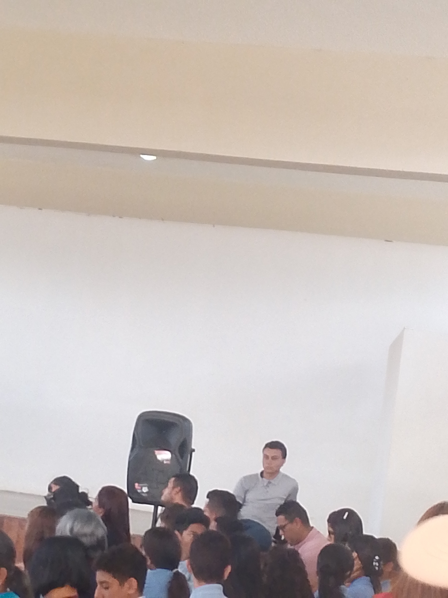 The image appears to show a group of people, possibly an audience, gathered in an indoor space with a large white wall behind them. A sound speaker stands to the left of the crowd. A man wearing a light grey shirt stands at the edge of the crowd, and other adults can be seen among what may be children. The setting is a large, possibly public space with a visible ceiling structure. There is no information in the image to identify if this location is in Juan Griego, Venezuela.