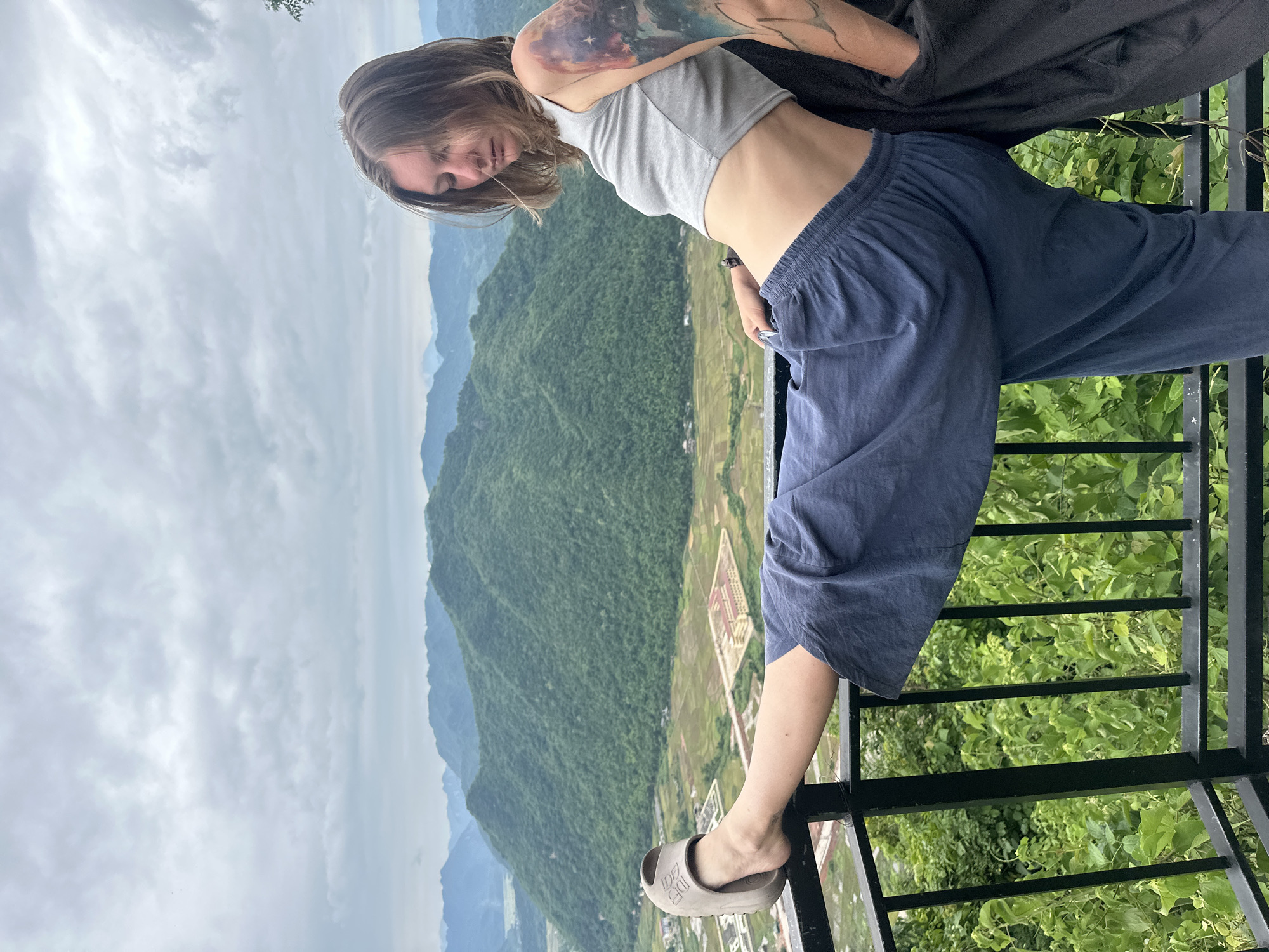 A young woman leans against a black metal railing with her legs extended, one foot resting on the railing. She is wearing a grey crop top and loose, dark blue pants. Her hair is light brown and falls over her shoulders. A large tattoo covers her left arm and shoulder. She is looking down with her eyes closed or partially closed.
Behind her, a lush, green valley stretches out, dotted with rice paddies and some buildings. Rolling, tree-covered mountains rise in the distance under a cloudy sky. This viewpoint appears to be from a high elevation, offering a panoramic view of the Mai Chau valley in Vietnam. The scene conveys a sense of relaxation and appreciation for the natural scenery.