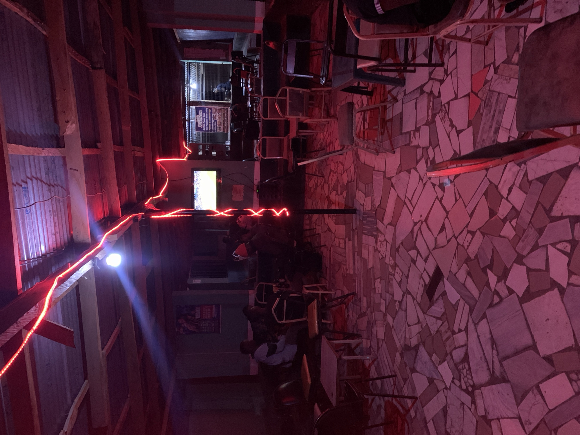 The image depicts an indoor space in New Stadium, Nigeria, illuminated by red LED strip lights. A group of people are seated watching a television screen. The room has a tiled floor with a mosaic pattern and a rudimentary wooden structure for a ceiling. Chairs and tables are scattered throughout the space. A sign can be seen in the background.