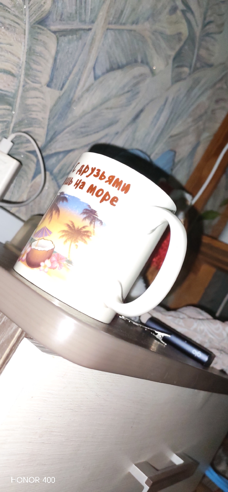 The image shows an indoor setting, likely a room, featuring a white mug placed on a light-colored wooden surface, possibly a nightstand or dresser. The perspective is tilted, looking slightly upwards.
The mug is the central subject, adorned with a colorful tropical scene depicting palm trees silhouetted against a sunset, a coconut drink with an umbrella, and pink hibiscus flowers. Above this design, there is text in Russian which reads: "С друзьями ночь на море," translating to "With friends, night at the sea."
To the left of the mug, a white power adapter or charger is visible near a wall. The wall is covered with wallpaper featuring a light blue and white pattern resembling large, stylized leaves or foliage, possibly with a subtle glittery texture.
On the wooden surface to the right of the mug, a dark blue or black cylindrical electronic device, possibly a vape pen, with "glo" written on its side, is lying next to a small, silver-colored clip. Below the main surface, a drawer of the furniture piece is visible, with a modern, brown and white handle.
The overall lighting suggests an indoor setting, possibly evening or artificial light. In the bottom left corner of the image, the watermark "HONOR 400" is visible, indicating the device used to capture the photo. No people are present, and no specific event or activity is depicted other than the static arrangement of objects.