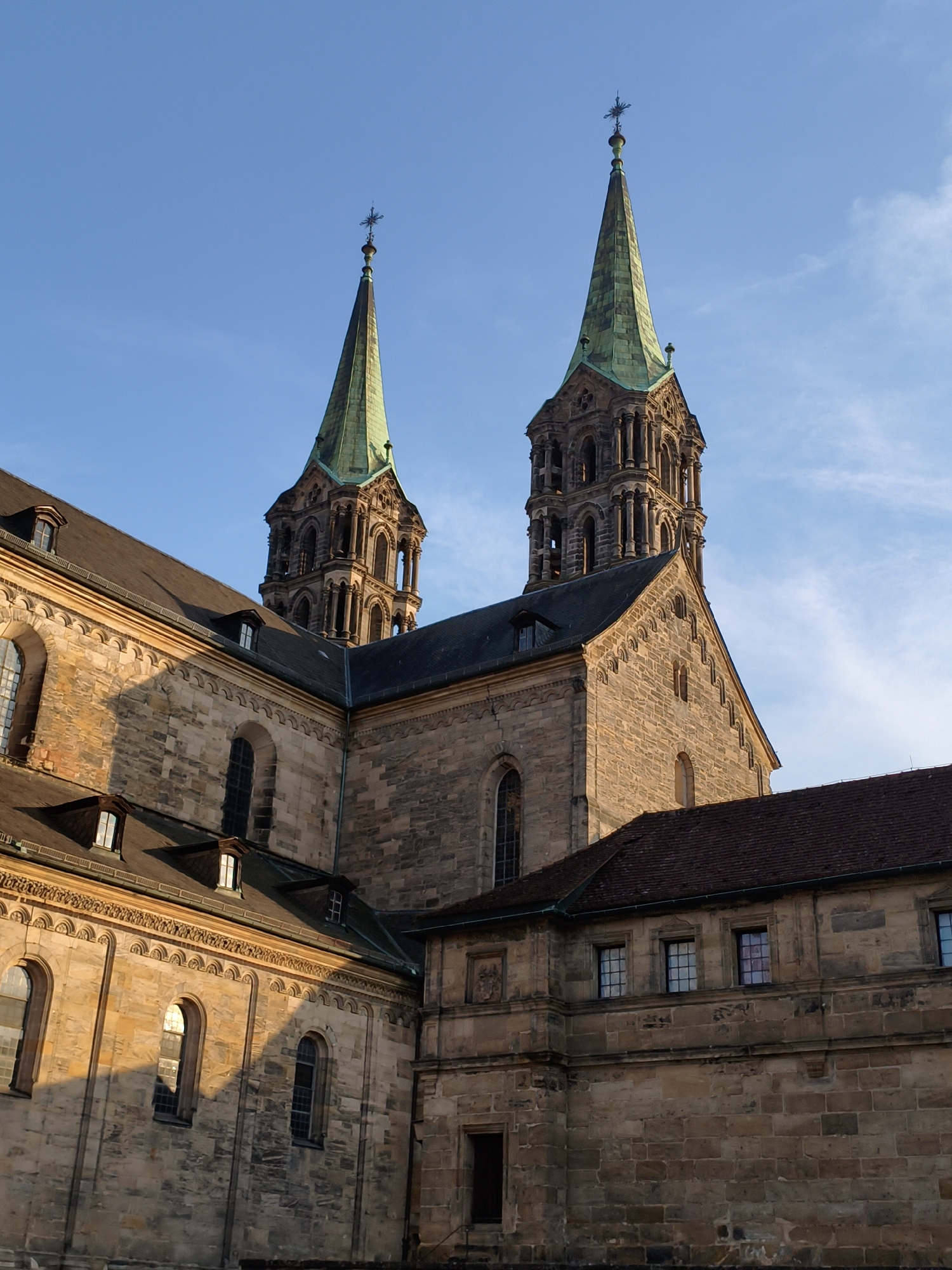 The image captures a detail of Bamberg Cathedral (Bamberger Dom) in Bamberg, Germany, under a clear blue sky with sparse clouds. The cathedral features two prominent spires, one with a distinctive green copper roof, topped with crosses. The building is constructed from light-colored stone, with intricate architectural details, including arched windows and ornate stonework. The sunlight casts long shadows across the facade, highlighting the texture of the stone and the depth of the architectural elements. The perspective emphasizes the grandeur and historical significance of the structure. There are no people or animals visible in the image, and no actions are taking place. The environment is solely the cathedral and the sky.