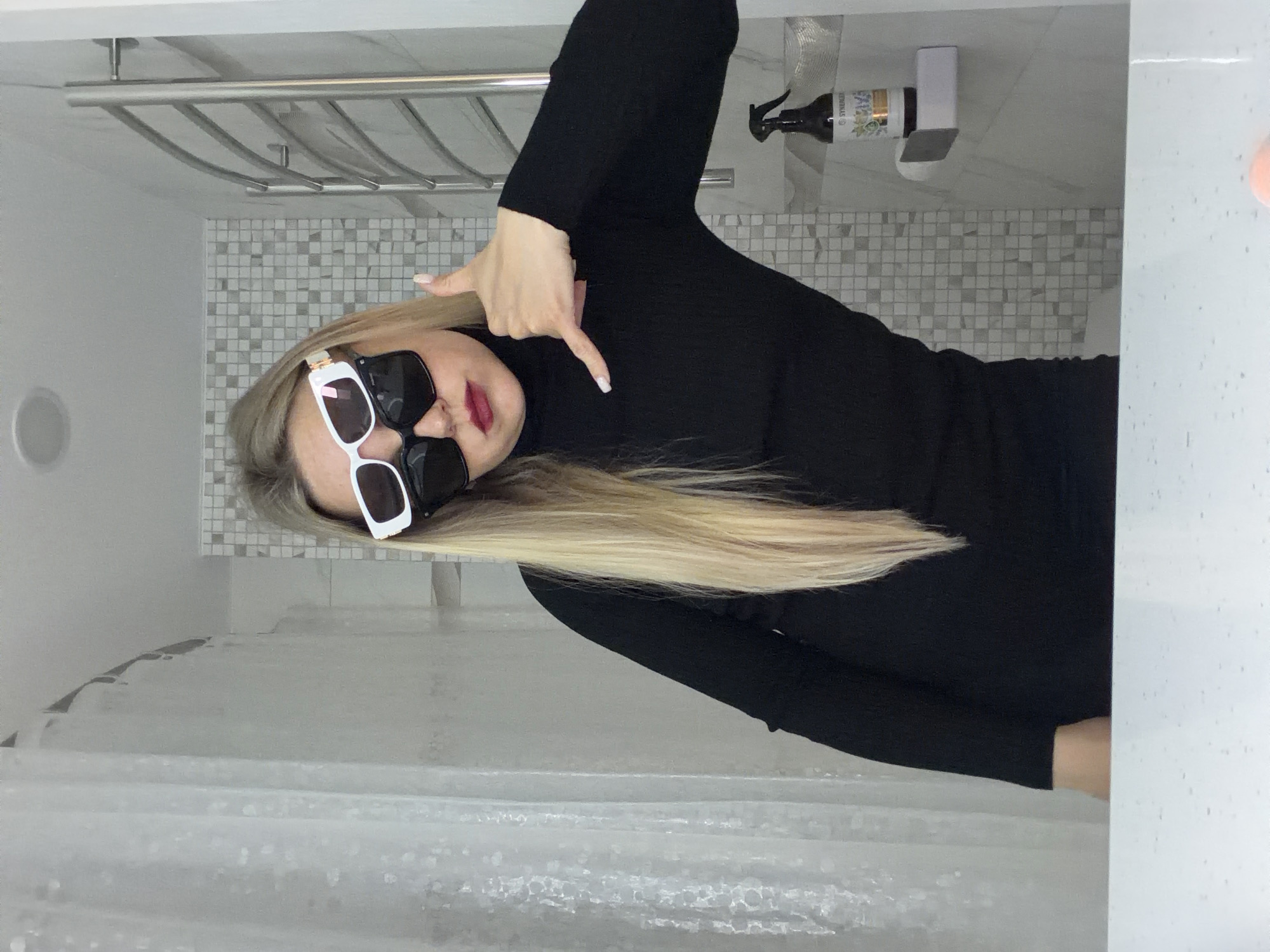 The image captures a young woman posing in what appears to be a bathroom in Votkinsk, Russia. The woman, with long, straight blonde hair, is wearing a black long-sleeved top and large, stylish white-framed sunglasses with dark lenses. Her left hand is raised to shoulder height, forming a "finger gun" gesture and pointing downwards. The image is rotated 90 degrees clockwise, presenting her horizontally across the frame.
The background elements suggest a modern bathroom. Behind her is a tiled wall featuring small, light and dark grey square tiles arranged in a mosaic pattern. Above this, a metallic towel rack is visible. To the right of the woman, a white countertop or shelf holds a black spray bottle with a label showing "SYNERGETIC" and "LAVENDER." A small white rectangular object rests beside the bottle. On the left side of the frame, a translucent, textured white shower curtain hangs. The woman's nails are neatly manicured. Her expression is mostly obscured by her sunglasses, but her pose conveys a confident or playful attitude. The lighting appears to be artificial, typical for an indoor setting, with no indication of the time of day.