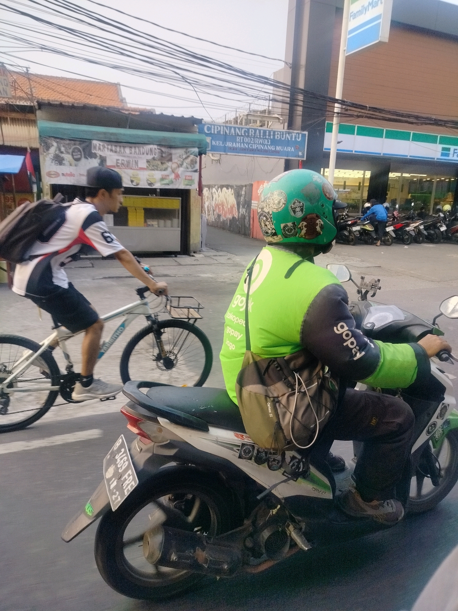In East Jakarta, Indonesia, on a street, two individuals are engaged in transit. On the left, a man with a black baseball cap rides a white bicycle. He wears a white and black shirt with red accents and black shorts, and a dark backpack is slung over his shoulders. In the foreground, a person on a scooter wears a bright green jacket with dark sleeves, decorated with various stickers. The jacket has "gojek" and "gopay" printed on it. The scooter rider also wears dark pants and a backpack. The street is paved, and buildings with awnings and signs line the background. One sign reads "MARTABAK BANDUNG ERWIN" and another, a blue street sign, indicates "CIPINANG BALI BUNTU". The sky is overcast, and electrical wires are visible overhead. The scene captures everyday street activity, with a focus on the two individuals and their modes of transportation.