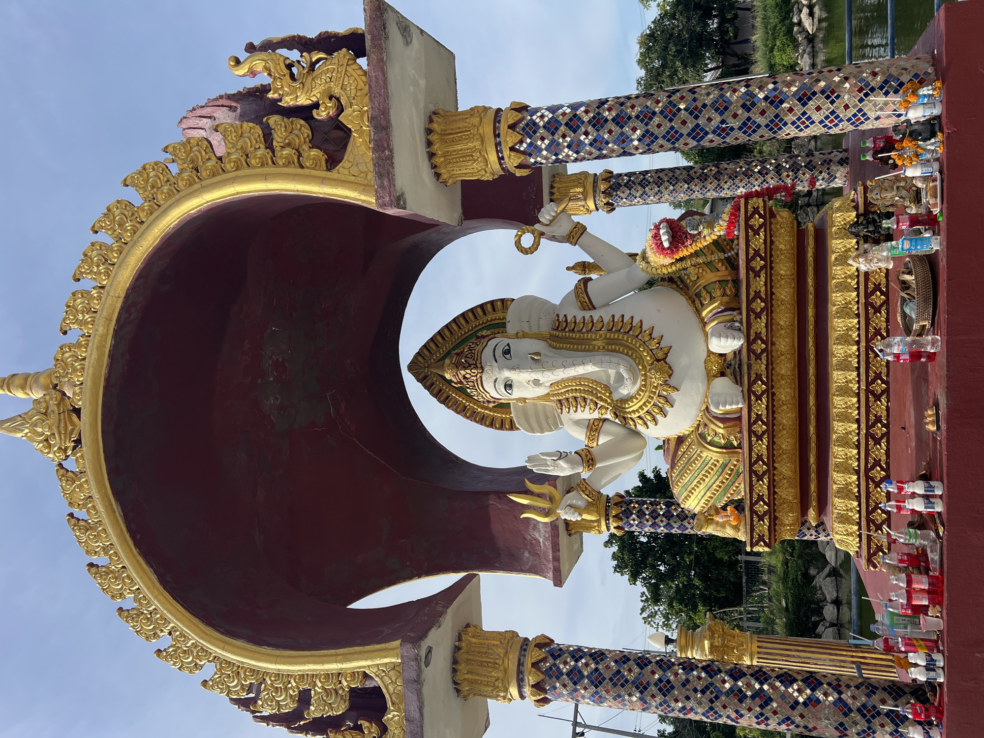 In Baan Plai Laem, Thailand, a large, white statue of the Hindu deity Ganesha is seated under a red and gold archway. Ganesha, with his characteristic elephant head and multiple arms, is adorned with intricate golden jewelry and a colorful floral garland. The statue is positioned on a raised, ornate platform decorated with gold and red patterns. In front of the platform, a red altar is laden with various offerings, including plastic bottles of drinks, small statues of deities, and floral arrangements. The altar is flanked by two cylindrical pillars covered in colorful mosaic tiles. Behind the altar, a body of water, likely a pond or canal, is visible, with trees and vegetation lining its banks. The sky above is a clear blue, with some clouds present. The overall scene depicts a religious shrine dedicated to Ganesha, set in an outdoor environment in Thailand.