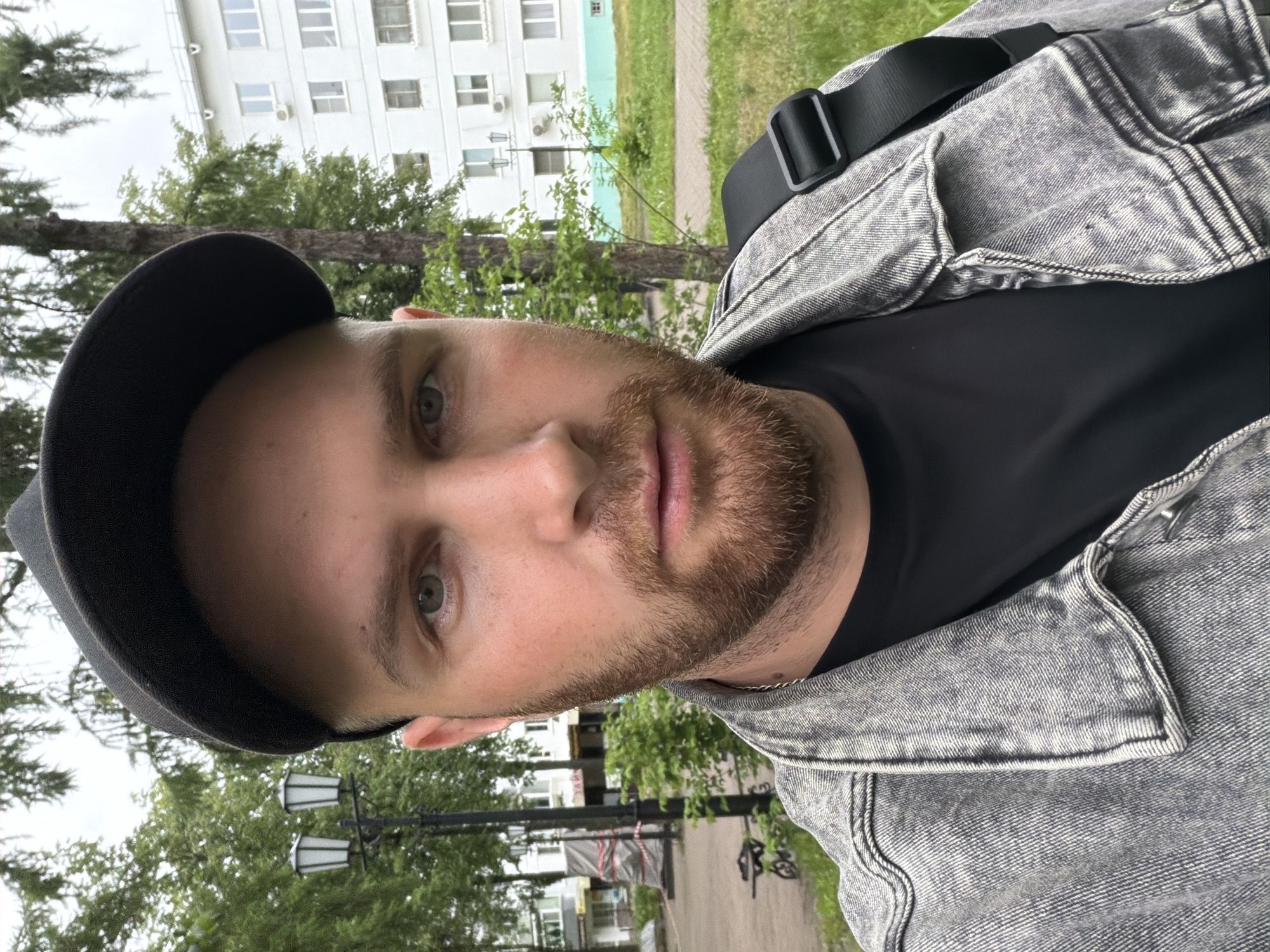 The image captures a male individual, shown from the chest up, facing directly forward in an outdoor urban environment within Chaikovsky, Russia. The subject wears a black baseball cap, a black t-shirt, and a grey acid-wash denim jacket. He has a reddish-brown beard and mustache, and light-colored eyes. A black strap, consistent with a shoulder bag, is visible over his right shoulder. The background features a paved path, green trees, and dense foliage under an overcast sky. To the left, a black street lamp with two lanterns is visible, along with a partial view of a dark metal bench. Further in the distance on the left, a large object covered in red and white tape is partially discernible, flanked by additional buildings. To the right, a multi-story white residential building with multiple windows and balconies is prominent, featuring an external air conditioning unit and a light blue stripe along its lower facade. A second, simpler street lamp stands near this building.