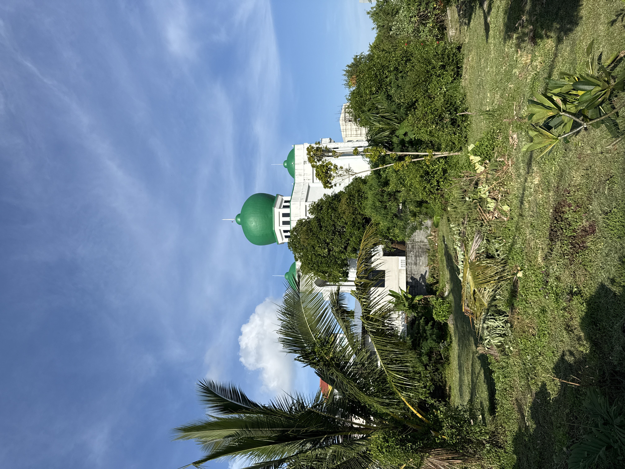 The image shows a mosque with green domes, a tall palm tree in the foreground, and lush green vegetation. The sky is blue with scattered clouds. The scene appears to be outdoors, likely in a tropical or subtropical environment given the presence of the palm tree and dense foliage. The architectural style of the building suggests it could be a place of worship, possibly a mosque due to the prominent domes. The overall impression is one of a serene and sunny day in a well-maintained area. The location is identified as Phuket City Municipality, Thailand.
