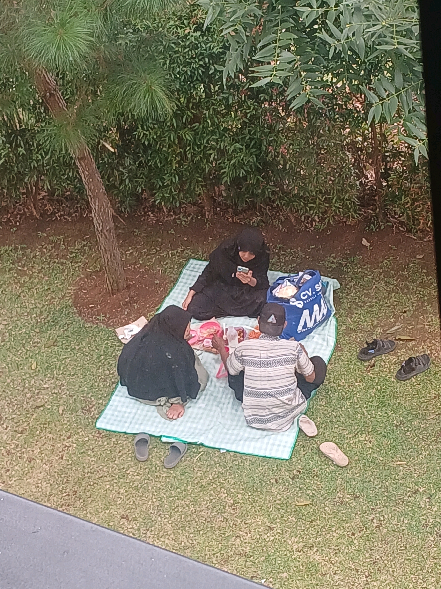 The image captures an aerial view of a picnic scene in Arjasari, Indonesia. Three individuals are seated on a light blue and white checkered blanket spread on a grassy area. One person, dressed in black Islamic attire, is looking at a mobile phone. Another individual, also in black Islamic attire, is positioned to the left and appears to be interacting with food on a tray. A third person, a male wearing a striped shirt and a dark cap, is seated to the right, facing away from the camera. A large blue tote bag with "CV. SA" visible on it rests near the center of the blanket. Two pairs of sandals are placed on the grass near the blanket, one pair of black sandals and another pair of lighter-colored sandals. The background consists of lush green foliage and the trunk of a tree.