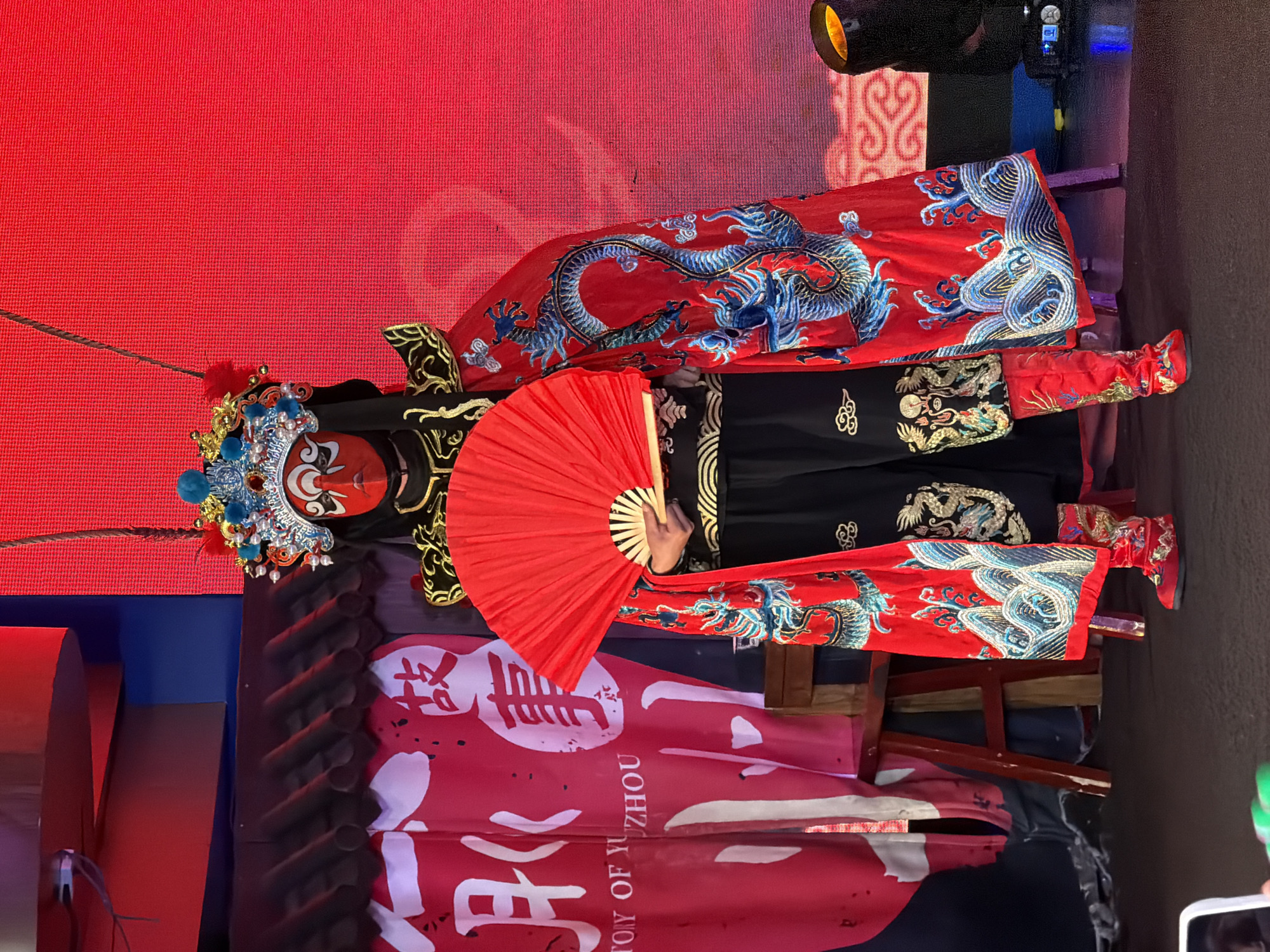 A performer in traditional Chinese opera costume stands on a stage, holding a red fan. The costume is elaborate, featuring a red mask with stylized facial features, a decorative headdress adorned with blue and red pompoms and beads, and a cape or robe with detailed blue dragon embroidery on a red background. The performer wears black pants with gold embellishments and red boots also decorated with gold patterns. In the background, red banners with Chinese characters and a stage with traditional Chinese architecture details are visible. A red LED screen forms the backdrop. The stage floor is dark, and theatrical lighting equipment is present on the right side of the image. The scene is set in Yuzhong District, China, likely at a cultural event or performance.