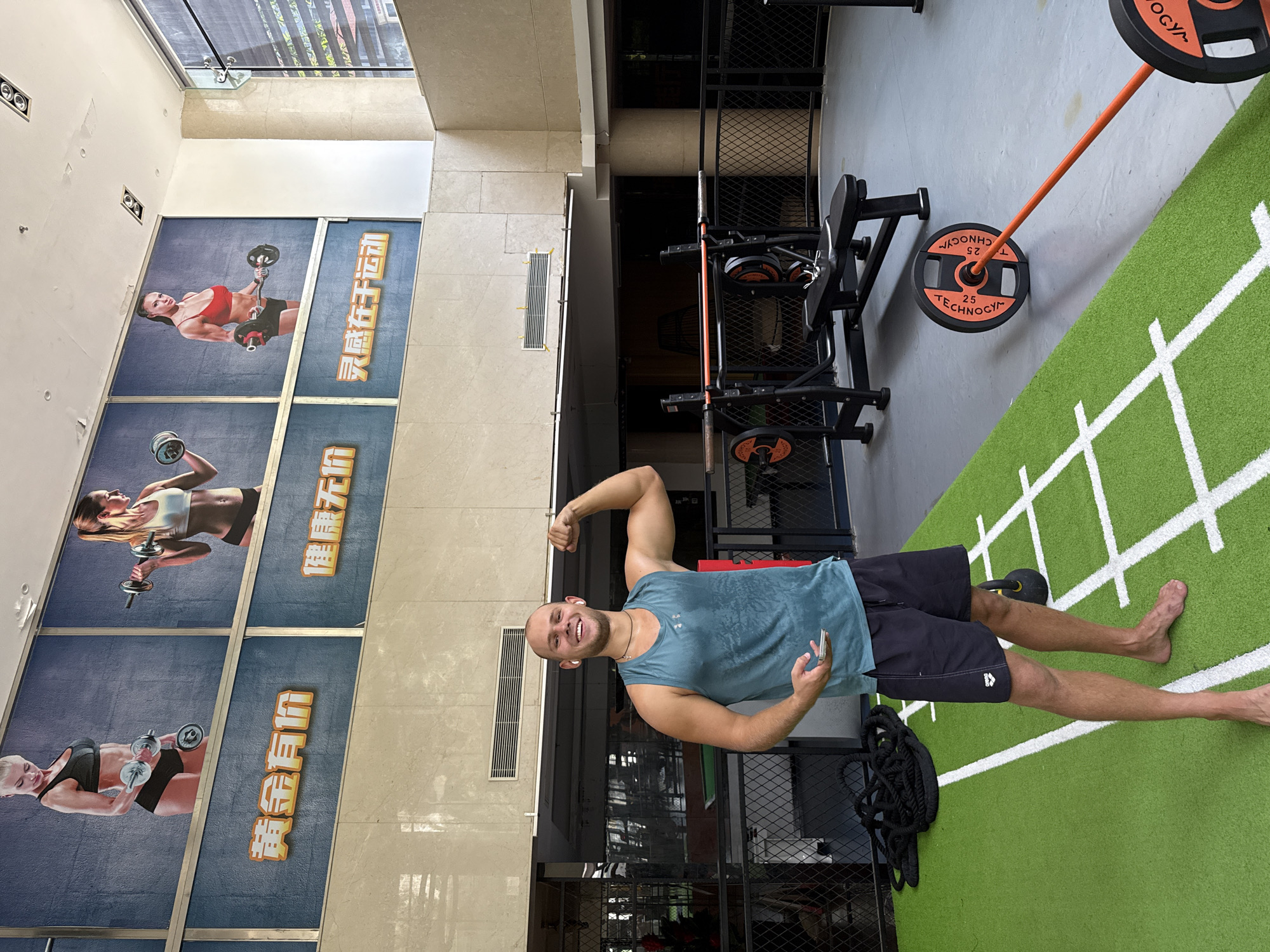 The image presents an indoor gym scene located in Haizhu District, China. A light-skinned adult male, bald with facial stubble, stands smiling towards the viewer. He wears a sweat-dampened blue-green sleeveless tank top, dark shorts with an "arena" logo on the left thigh, and white wireless earbuds. A dark necklace with a cross pendant is visible. He is barefoot, flexing his left bicep while holding a light-colored smartphone in his right hand. He is positioned on a green artificial turf section marked with white lines, typical of a workout area. To his immediate left on the turf, a coiled black battle rope is visible, and a black kettlebell is partially visible to his right.
Behind the man, the gym floor transitions to a grey surface, where a black weight bench with an integrated barbell rack stands. An orange barbell with two black and orange Technogym-branded 25kg weight plates is lying diagonally on the grey floor in the mid-ground. The gym space is delineated by black metal fencing.
Above the gym floor, a series of three large murals are mounted on an upper wall section. Each mural depicts a fit woman engaged in weightlifting. The leftmost mural shows a blonde woman with dumbbells, the central mural features a blonde woman with a single dumbbell, and the rightmost mural displays a dark-haired woman with a barbell. Below these murals, yellow-orange Chinese characters are displayed. From left to right, these translate to "Gold has a price," "Health is priceless," and "Inspiration comes from exercise." The upper walls are constructed from light-toned tiled surfaces, incorporating ventilation grilles and recessed lighting fixtures. A large window on the far right side of the image reveals reflections of exterior buildings.