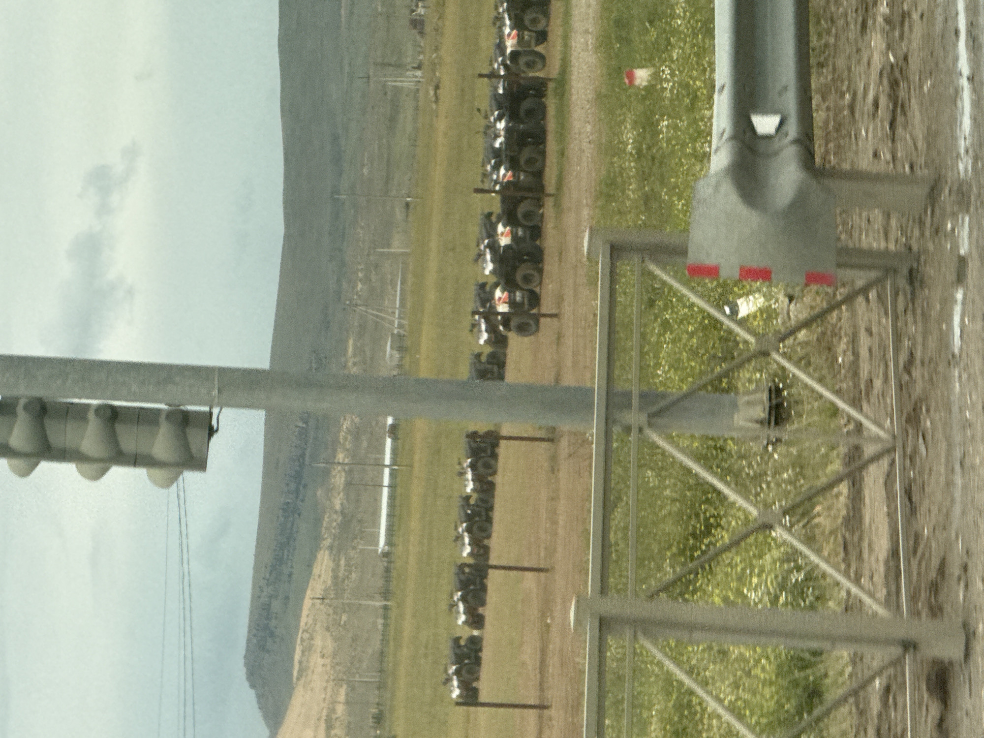 The image, oriented on its side, captures an outdoor scene at what appears to be an ATV staging or storage area, likely in Korkmaskala, Russia.
In the immediate foreground on the right, a large, dark gray metallic structure, possibly a gantry or support pillar, stands prominently. It features red rectangular markings near its base and a white rectangular opening higher up. This structure is integrated with a lighter gray metal framework exhibiting X-shaped cross-bracing.
Extending into the mid-ground, a long line of all-terrain vehicles (ATVs) is parked neatly on a dirt path adjacent to a grassy field. The ATVs are dark in color, with some displaying visible white or red accents. Behind this main row, another, less distinct line of ATVs suggests a larger collection of these vehicles.
To the left, another tall, slender metallic pole supports an overhead horizontal bar from which several cone-shaped objects, possibly loudspeakers or light fixtures, hang. Multiple thin cables run vertically downwards from this fixture. The ground in this area is covered in green grass interspersed with small white flowers.
The background reveals undulating hills or mountains beneath an overcast or partly cloudy sky. The scene depicts a daytime setting. There are no visible people or discernible text in the image.