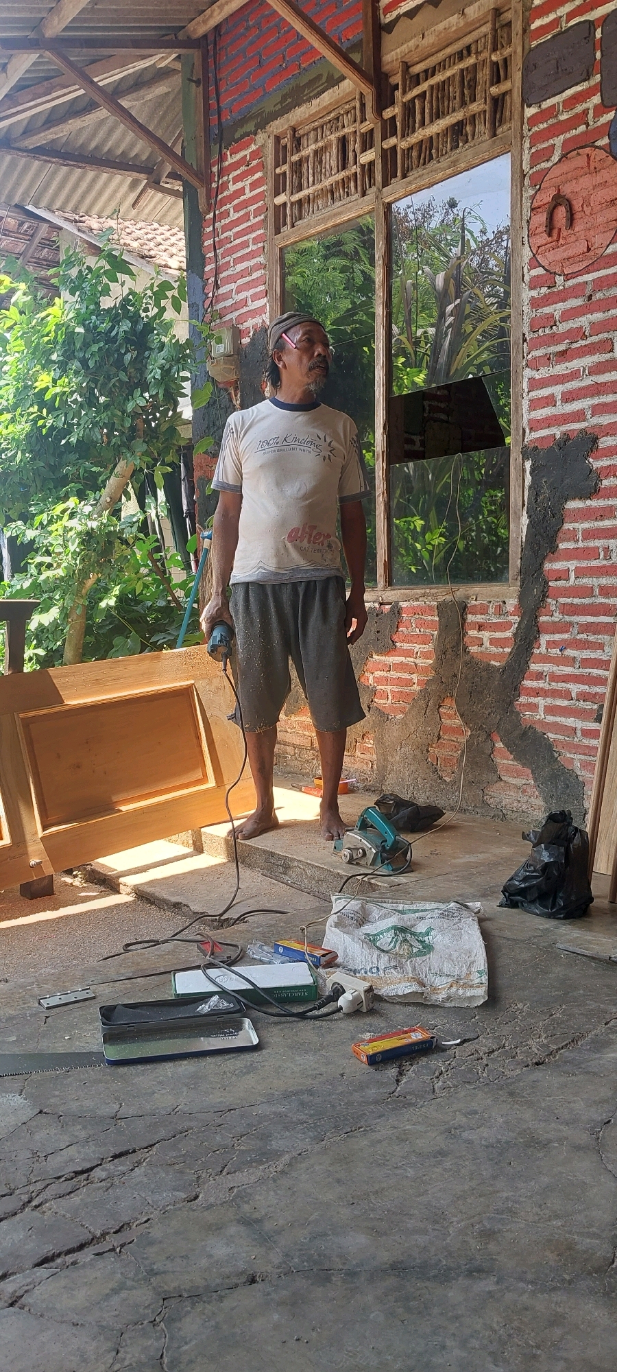 The image depicts an outdoor scene in Keling, Indonesia, likely a residential construction or renovation site. A middle-aged man with a mustache and wearing a light-colored t-shirt and dark shorts stands near a doorway or window frame. He is holding a power tool, possibly a drill. In front of him, a wooden door or frame lies on the ground, with sawdust scattered around.
Several tools and materials are present on the concrete floor, including a planer, electrical cords, a power strip, and what appear to be boxes of supplies and possibly some type of sealant or adhesive. A plastic bag and a black trash bag are also visible. The background shows a brick wall, some greenery from surrounding vegetation, and a corrugated roof. The overall impression is one of ongoing manual labor in a semi-finished outdoor space.