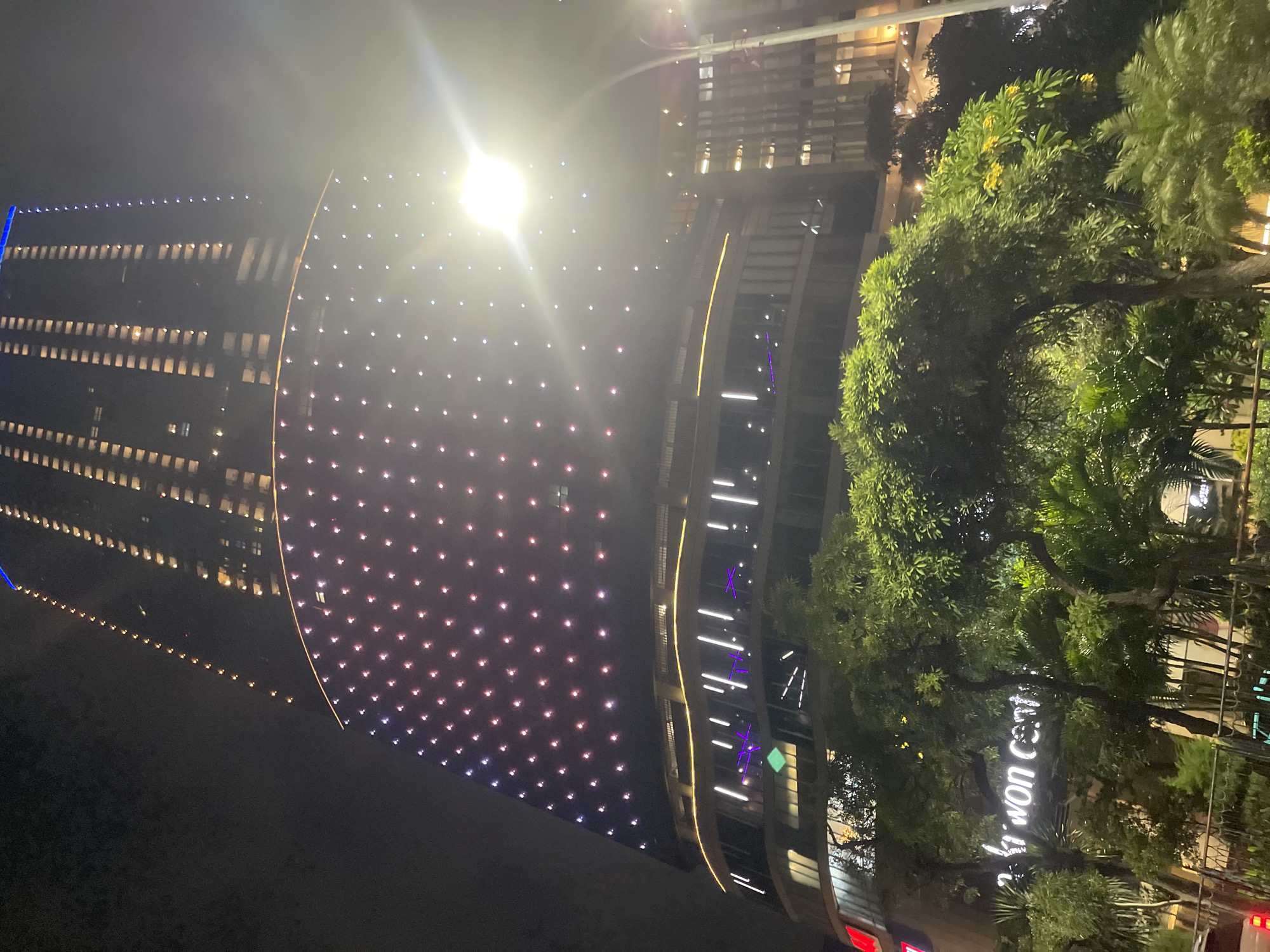 This is a nighttime photograph of a tall, modern skyscraper in Surabaya, Indonesia, illuminated with numerous small lights. The building features a curved lower section covered in a grid of pink and purple lights, with a brighter, more vertical upper section where individual windows are lit. A bright streetlamp is visible to the right, its light casting a glare across the upper portion of the building. In the foreground, lush green trees and foliage obscure the lower part of the building, with a neon sign reading "Ki won cent" visible through the leaves. The overall scene depicts a vibrant urban environment at night, likely a commercial or business district.